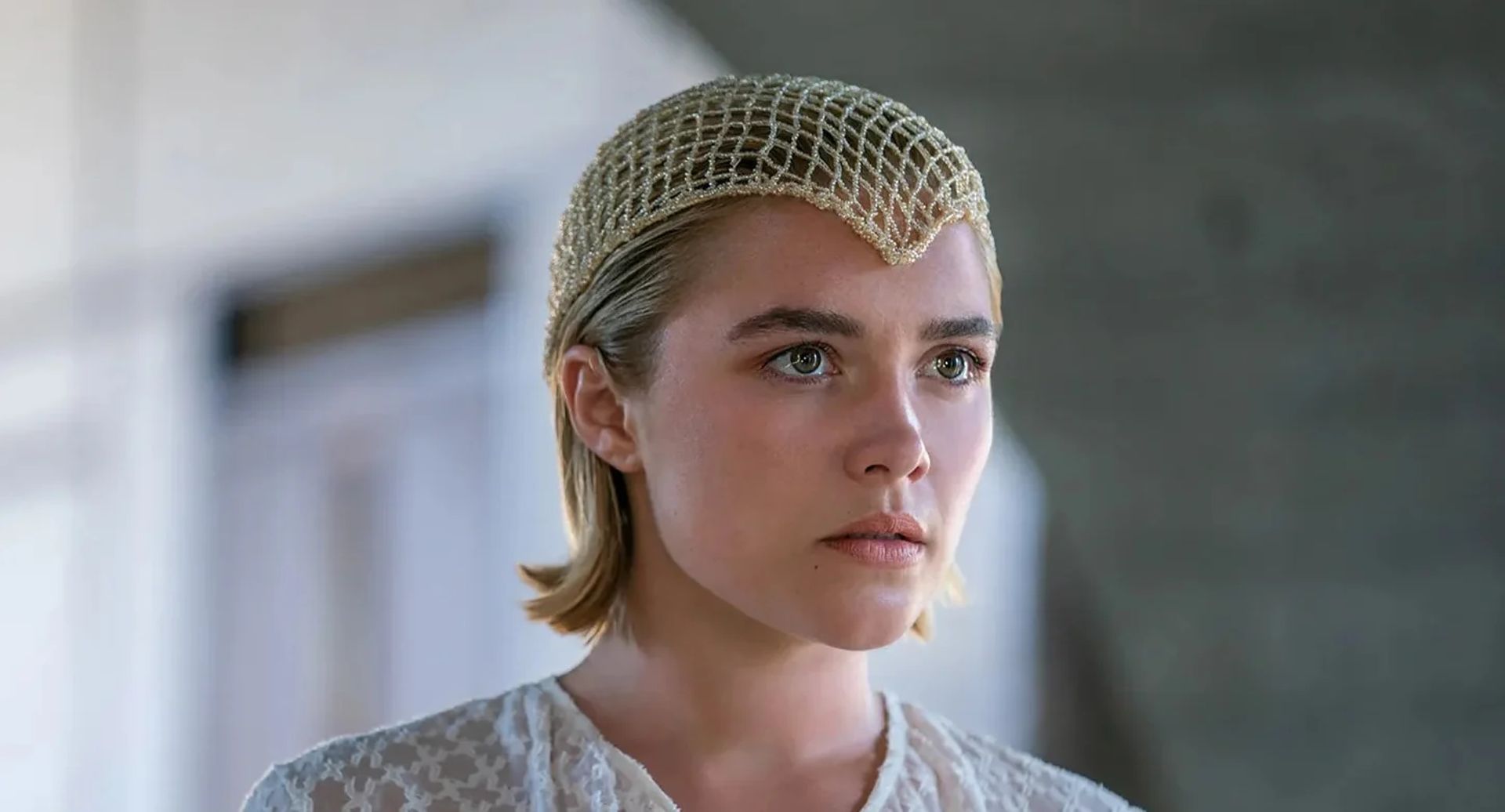A young woman with short blonde hair wears a delicate, gold mesh headpiece and a white lace top, looking intently ahead—her striking presence hints at one of the stars to watch 2026. The background is softly blurred with neutral tones.