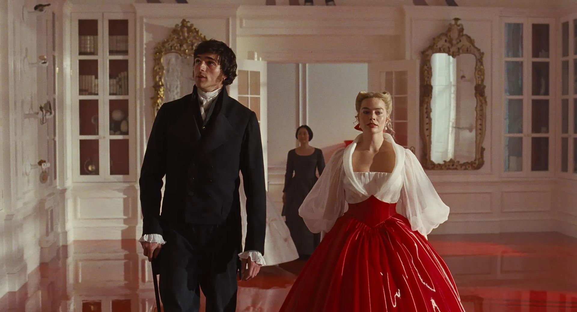 A man in a black suit and a woman in a striking red and white gown walk side by side in an ornate corridor with gold mirrors, while another woman stands in the background—capturing the elegance of the Stars to Watch 2026.