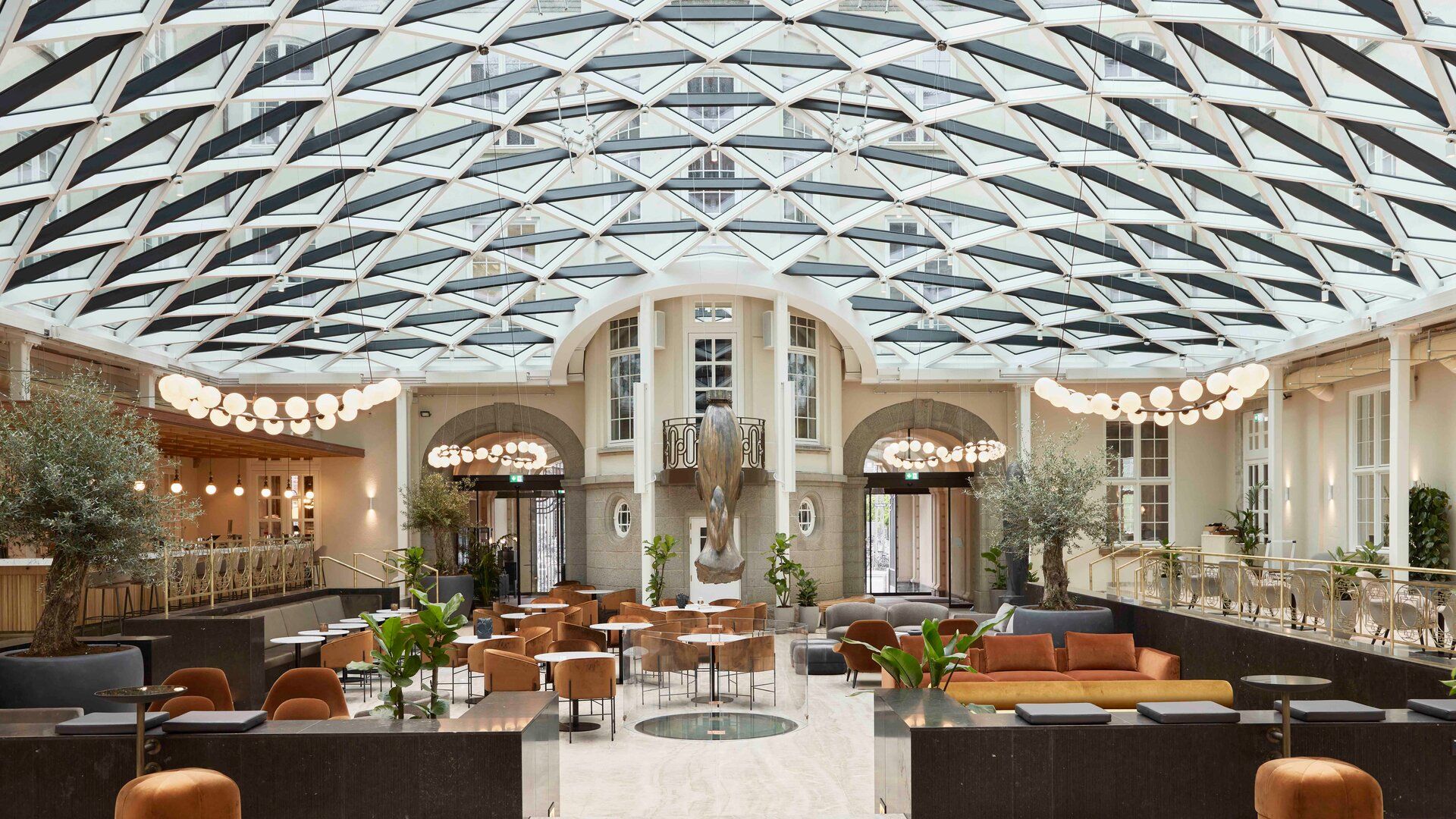 Modern hotel lobby at Villa Copenhagen with a glass-domed ceiling, stylish earth-toned seating, indoor trees, round lights, a grand staircase, and large windows—an inviting space to begin your travel 2026 adventure in Copenhagen.