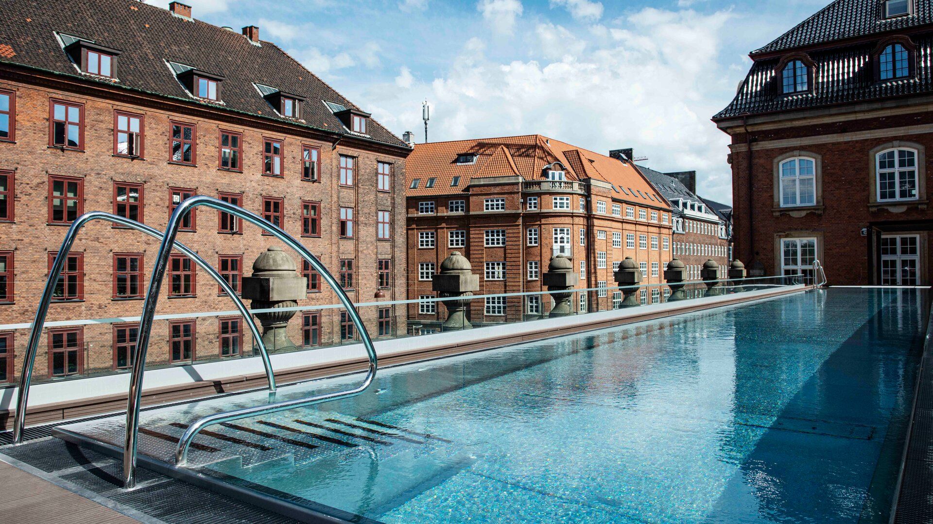 A rooftop swimming pool with metal handrails, surrounded by a glass barrier, overlooks Copenhagen’s historic red-brick buildings with gabled roofs—an inviting scene for your 2026 travel plans.