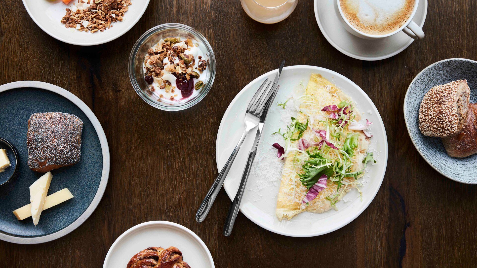 A breakfast spread at Villa Copenhagen features an omelette with greens, a seeded roll, cappuccino, yoghurt with granola, juice, pastry, cheese slices, and a chocolate muffin—perfect for fuelling your 2026 travel adventures in Copenhagen.