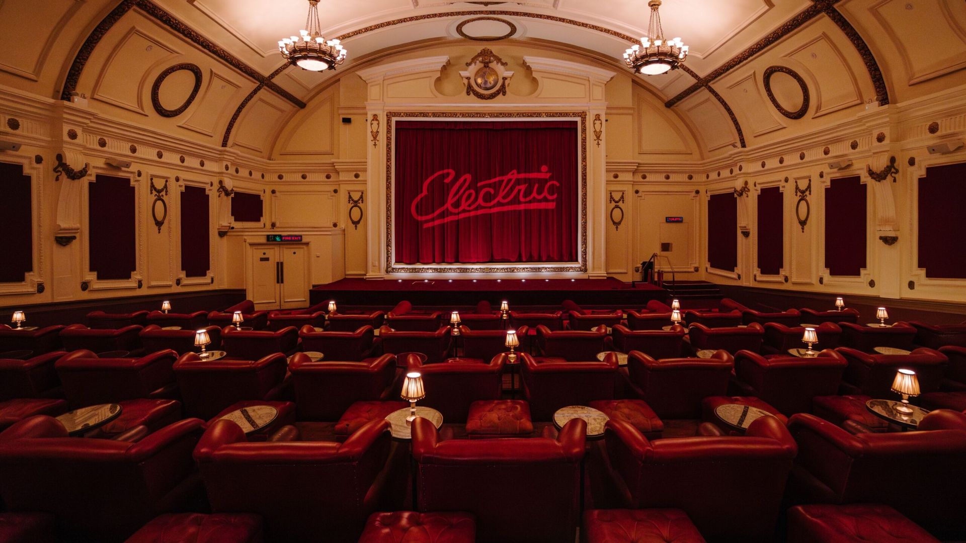 An ornate vintage cinema with red velvet seats and small tables, perfect for a rainy day in London, faces a stage with red curtains and the word Electric in neon. Elegant chandeliers hang from the high, detailed ceiling.