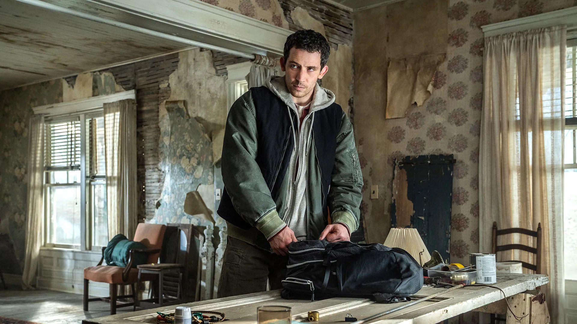 A man in a dark jacket stands at a worn table in a shabby, dimly lit room with peeling wallpaper and old furniture, packing items into a black bag—like a scene straight from the biggest films 2026 is set to deliver.