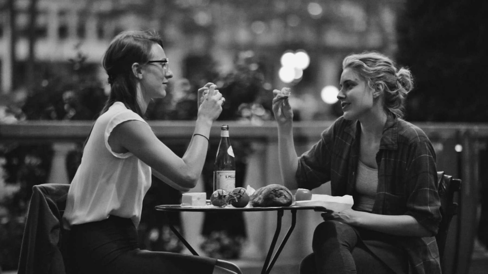 Two women sit at a small outdoor table, sharing food and drinks—in a scene straight out of the best romantic comedies. One wears glasses and a sleeveless top, the other a checked shirt; bread, fruit, and wine complete the vibrant romcom vibe.