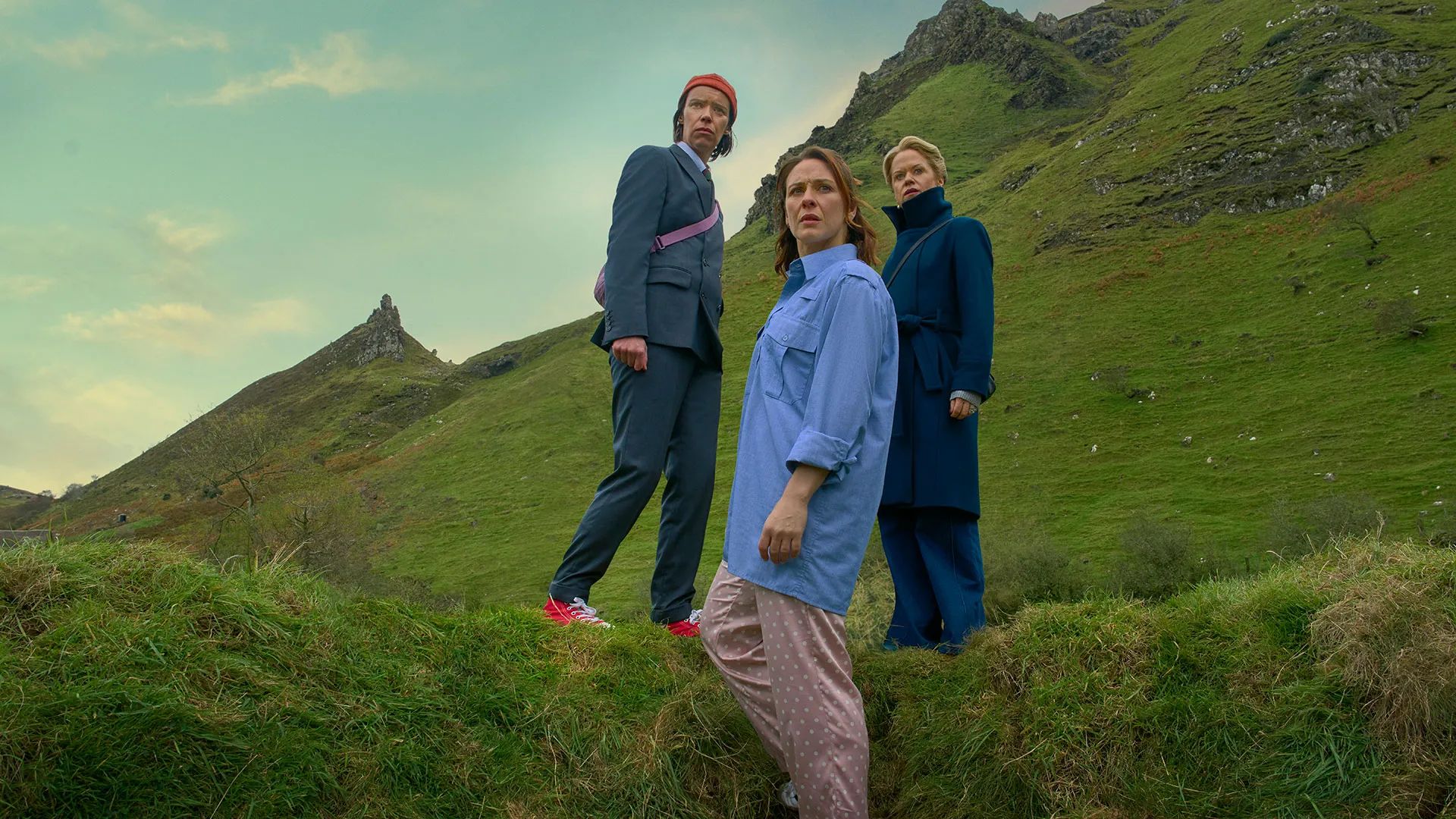 Three people stand on a grassy hill with rocky slopes behind them, dressed in casual, quirky clothing, looking intently towards the camera under a cloudy sky—wondering how to get to heaven from Belfast.