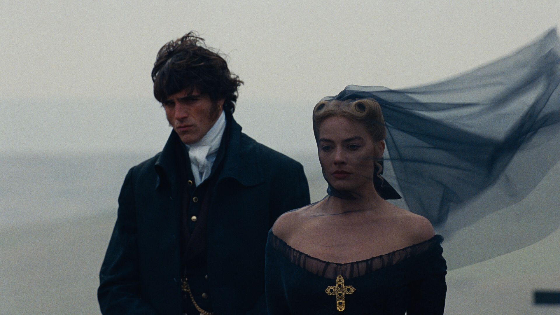 A sombre man and woman, reminiscent of Wuthering Heights, stand on a foggy, windswept beach. The woman, echoing Margot Robbie’s poise, wears a black dress with a gold cross and veil; the man stands behind her, looking solemn.