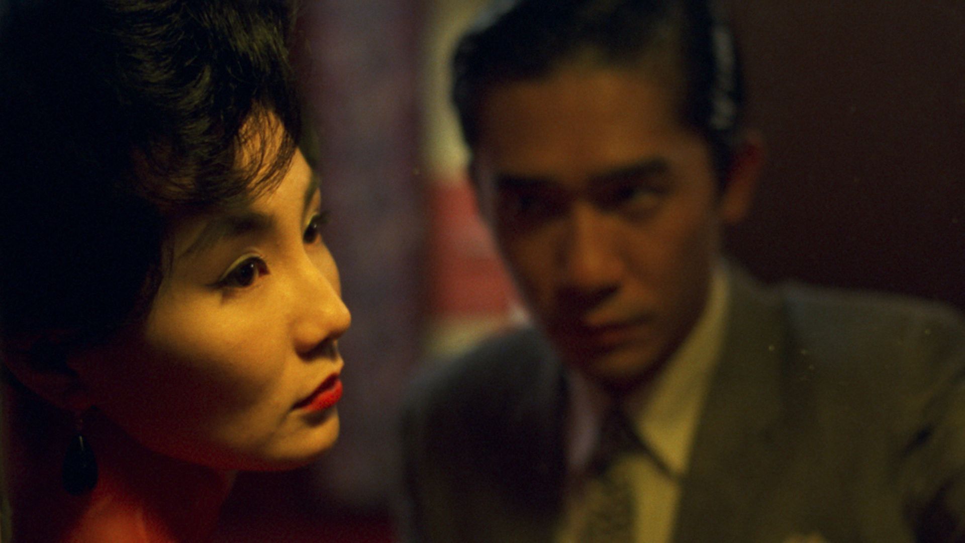 A woman with short dark hair and red lipstick gazes thoughtfully to the left, while a man in a suit sits behind her in soft focus, channelling the intimate, moody vibe often found in the best romantic comedies from a bloke’s perspective.