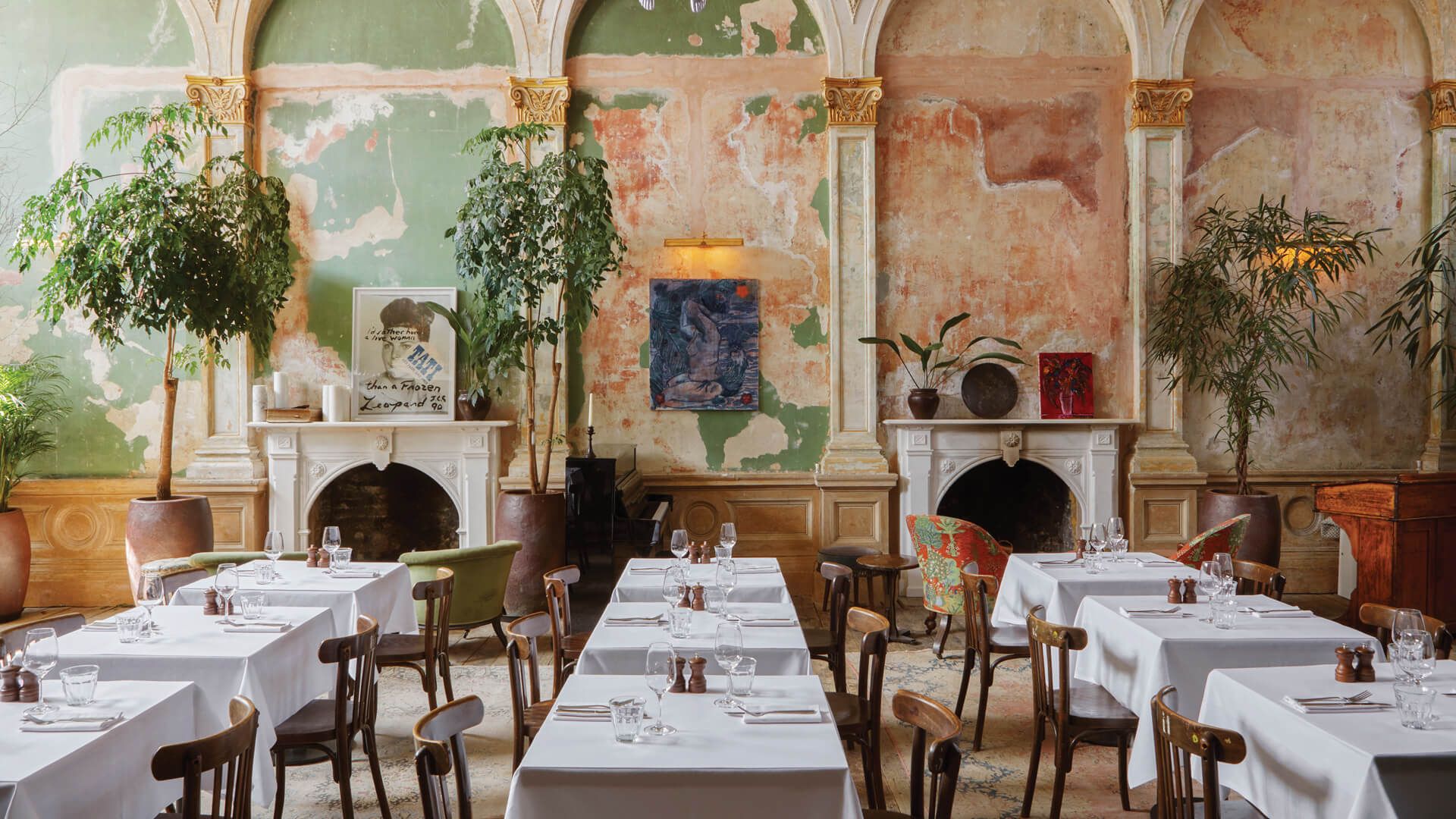 A stylish restaurant designed by Lucy Barlow, with white tablecloths, wooden chairs, potted plants, and two ornate fireplaces set against a vintage, peeling pastel wall with eclectic artwork. The atmosphere is elegant and rustic.