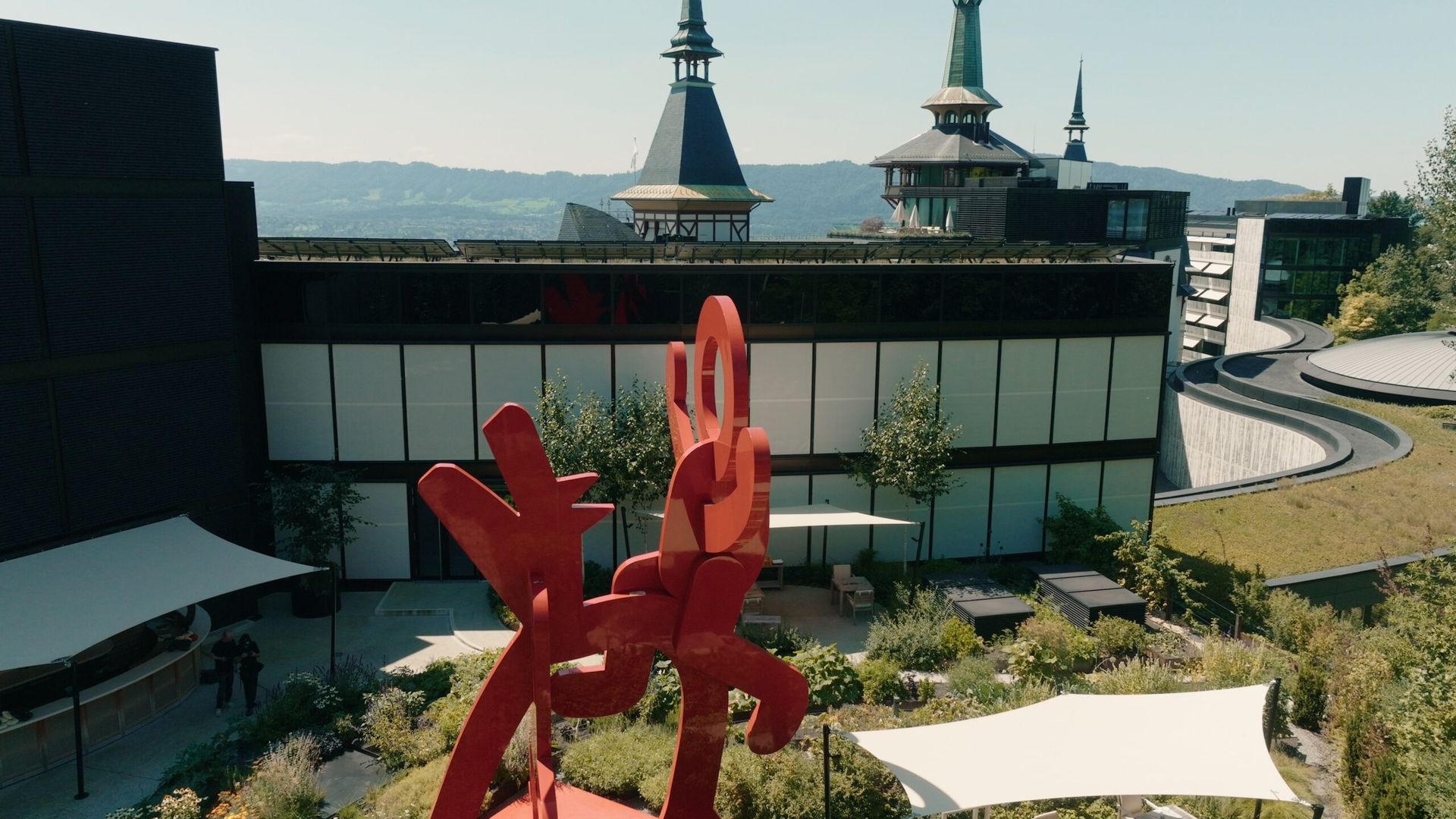 A large, abstract red sculpture stands in a landscaped courtyard at the Dolder Grand, a fairytale hotel in Zurich, surrounded by modern and historic buildings with towers and spires, under a clear sky.