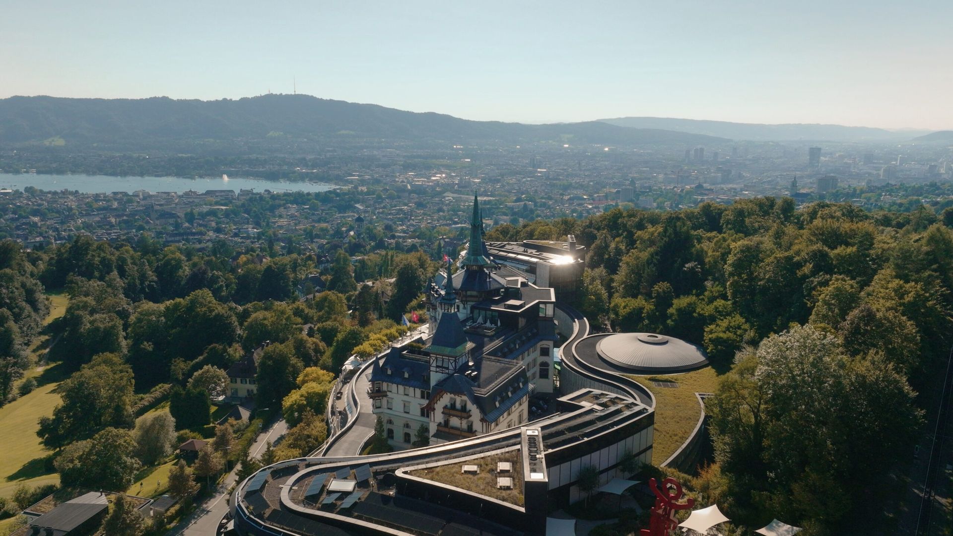 Aerial view of the fairytale hotel Dolder Grand, nestled among lush green trees in Zurich, with a cityscape, lake, and hills stretching beneath a clear sky.