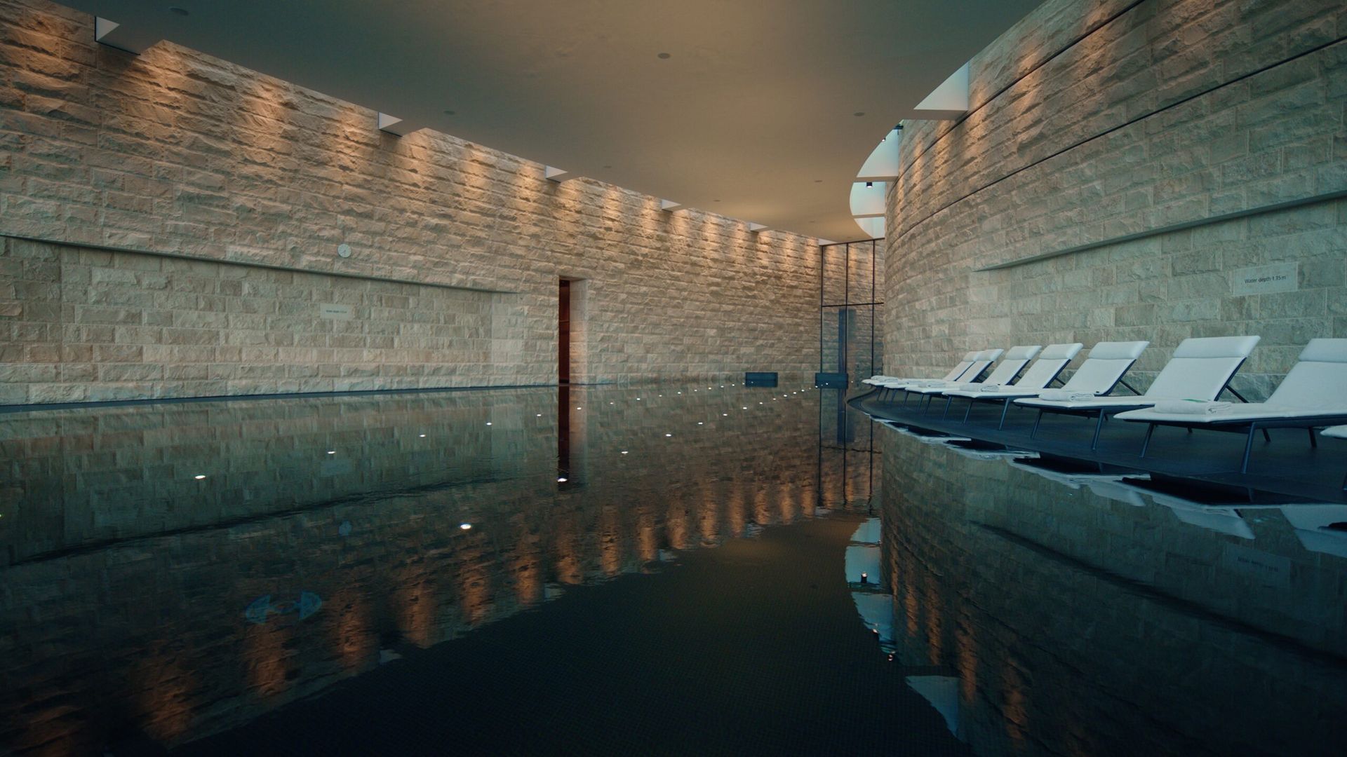 A modern indoor swimming pool at the Dolder Grand, Zurich’s fairytale hotel, features stone walls, a high ceiling, and recessed lighting. White loungers line one side as the shimmering water reflects the elegant surroundings.