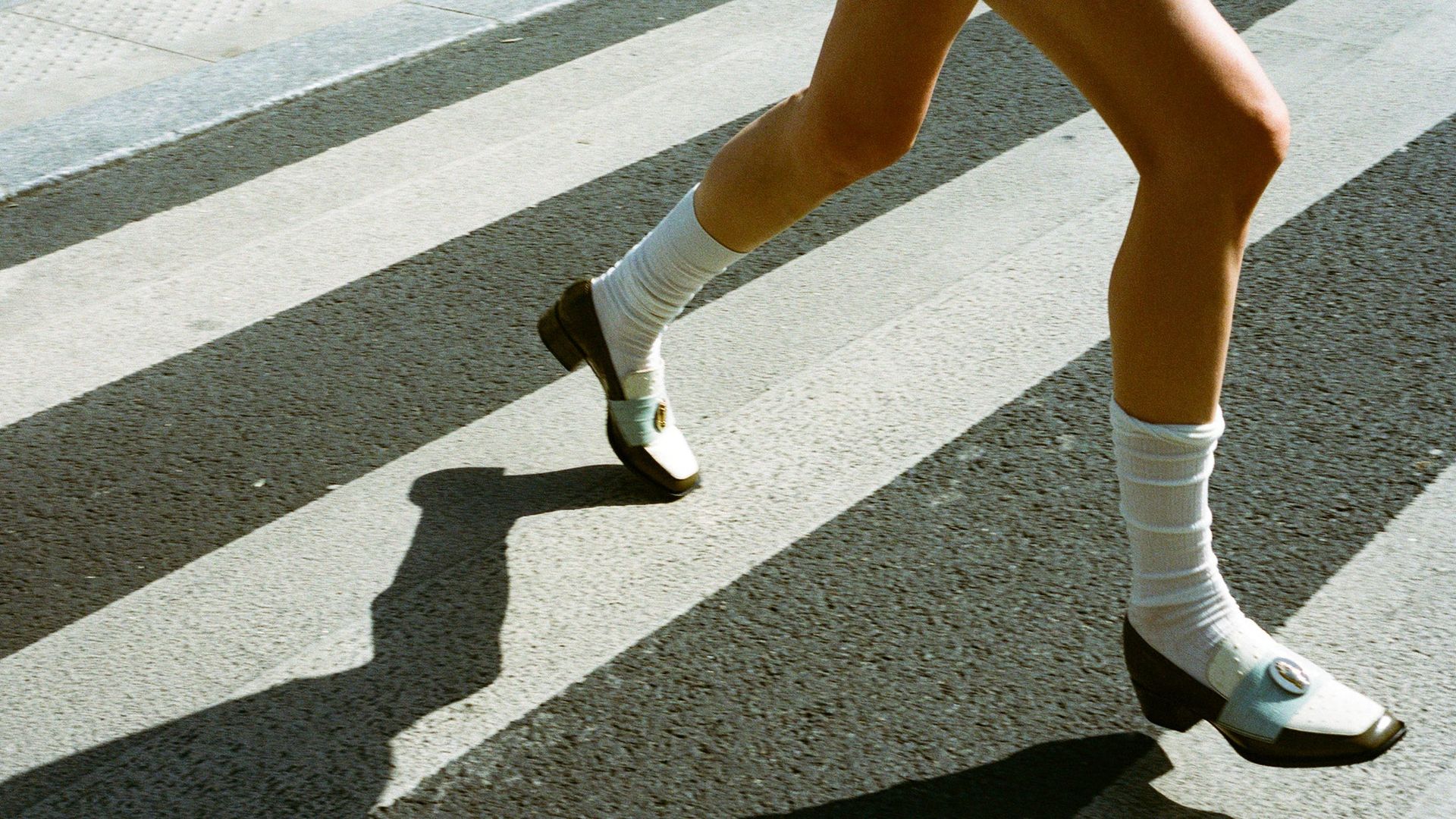 A person wearing white socks and black shoes from small fashion brands quickly walks across a zebra crossing, casting a strong shadow on the pavement.