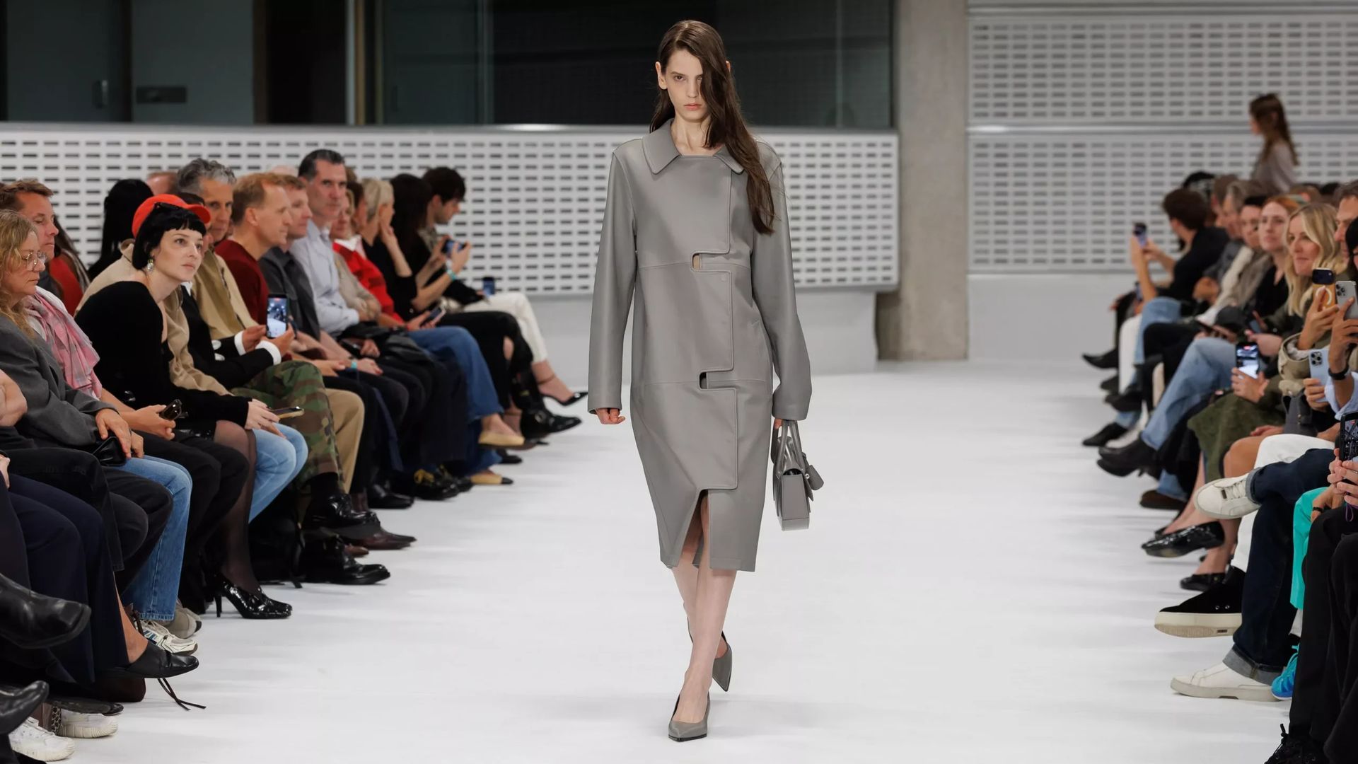 A model wearing a grey coat, carrying a matching handbag, and walking down a catwalk as an audience watches from both sides, captures the dynamic atmosphere of London Fashion Week.