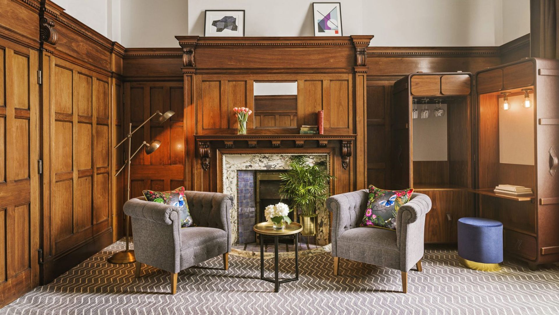 A cosy sitting area with two grey armchairs, a small round table, colourful pillows, and a fireplace with a wooden mantel—this stylish space could easily be featured in any Manchester Guide for contemporary décor inspiration.