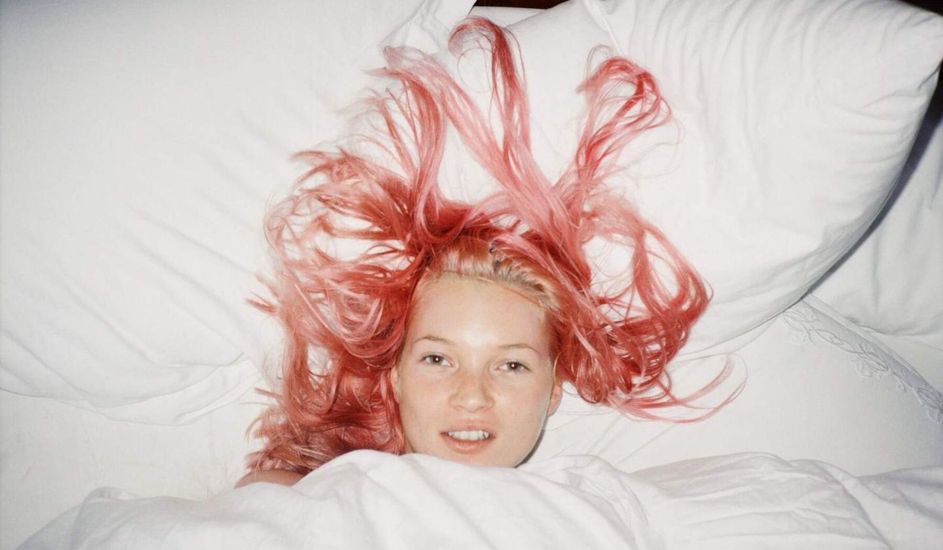 A person with long, pink hair lies in bed, partially covered by a white blanket and surrounded by white pillows, their striking hair spread out—reminiscent of scenes from Fashion Exhibitions.