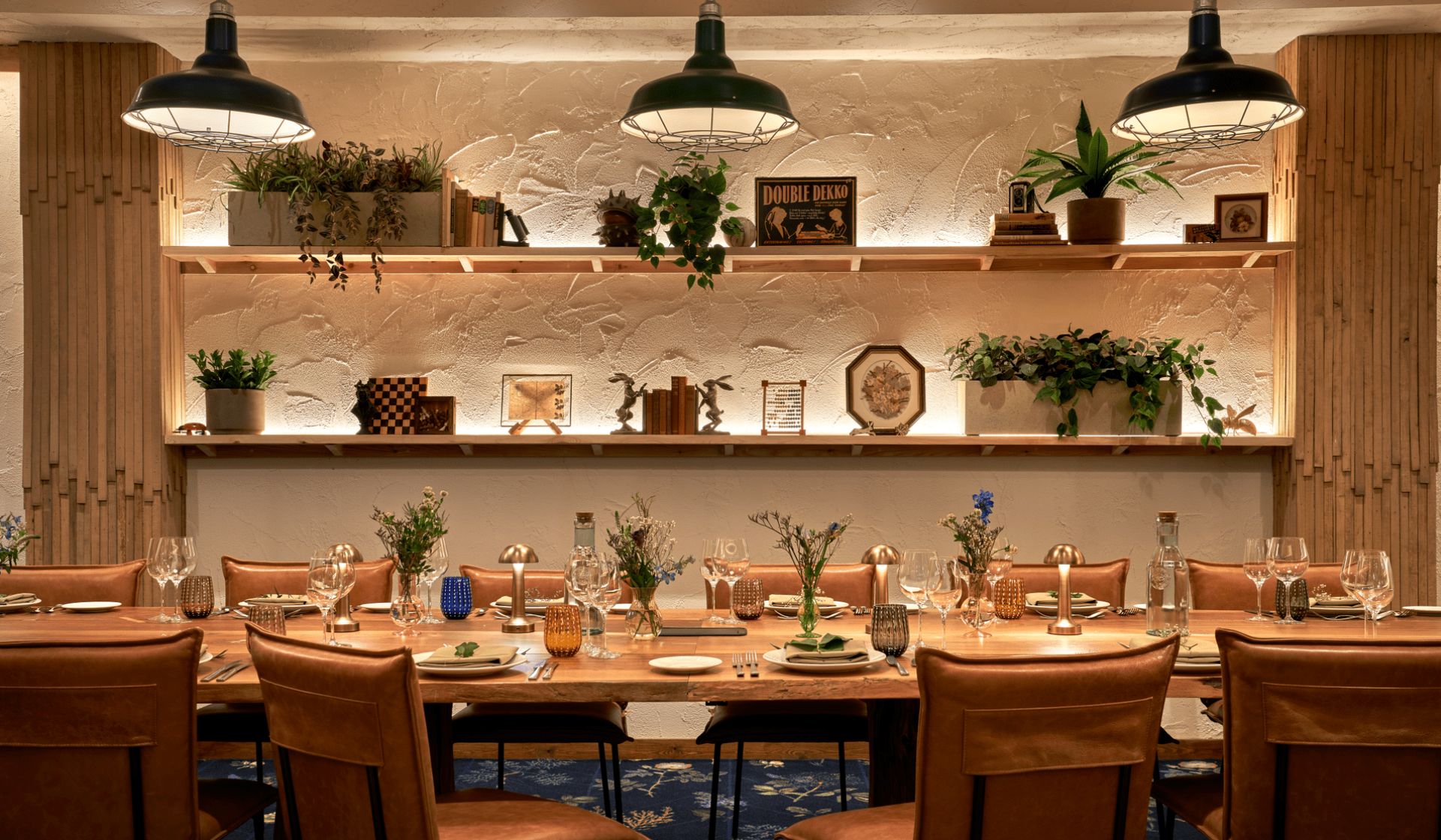 A warmly lit dining room with a long wooden table set for a meal, surrounded by tan leather chairs—an inviting spot to add to your Manchester guide for where to eat. Shelves display plants and books, whilst pendant lights hang from the ceiling.