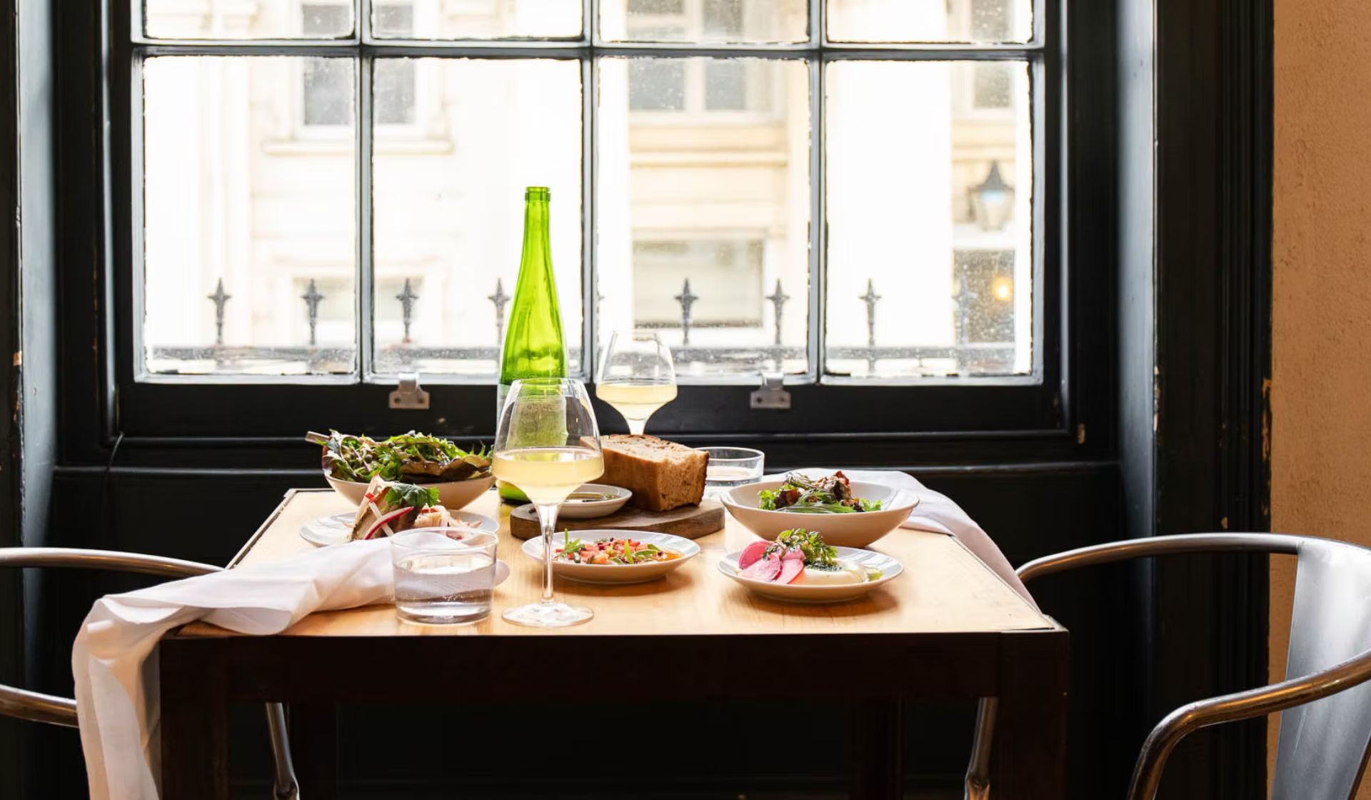 A wooden table set for two by a window in Brighton, featuring plates of food, white wine, bread, salad, and water with metal chairs—perfect for relaxing after exploring the best things to do in Brighton.