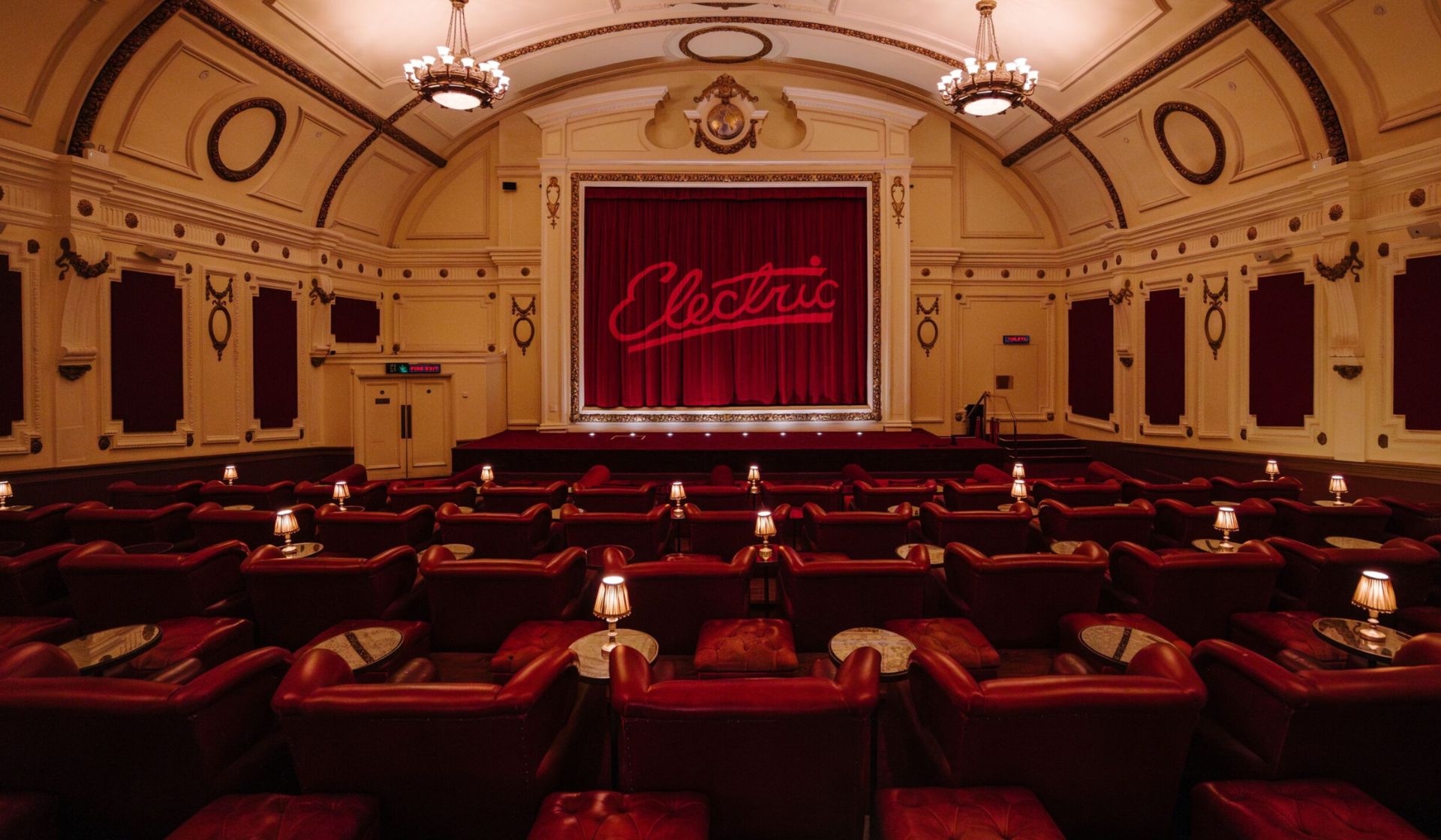A vintage cinema perfect for indoor activities, with plush red armchairs, small tables with lamps, and an ornate ceiling. The stage features a red curtain adorned with "Electric" in neon-style letters. The room is elegant and warmly lit.