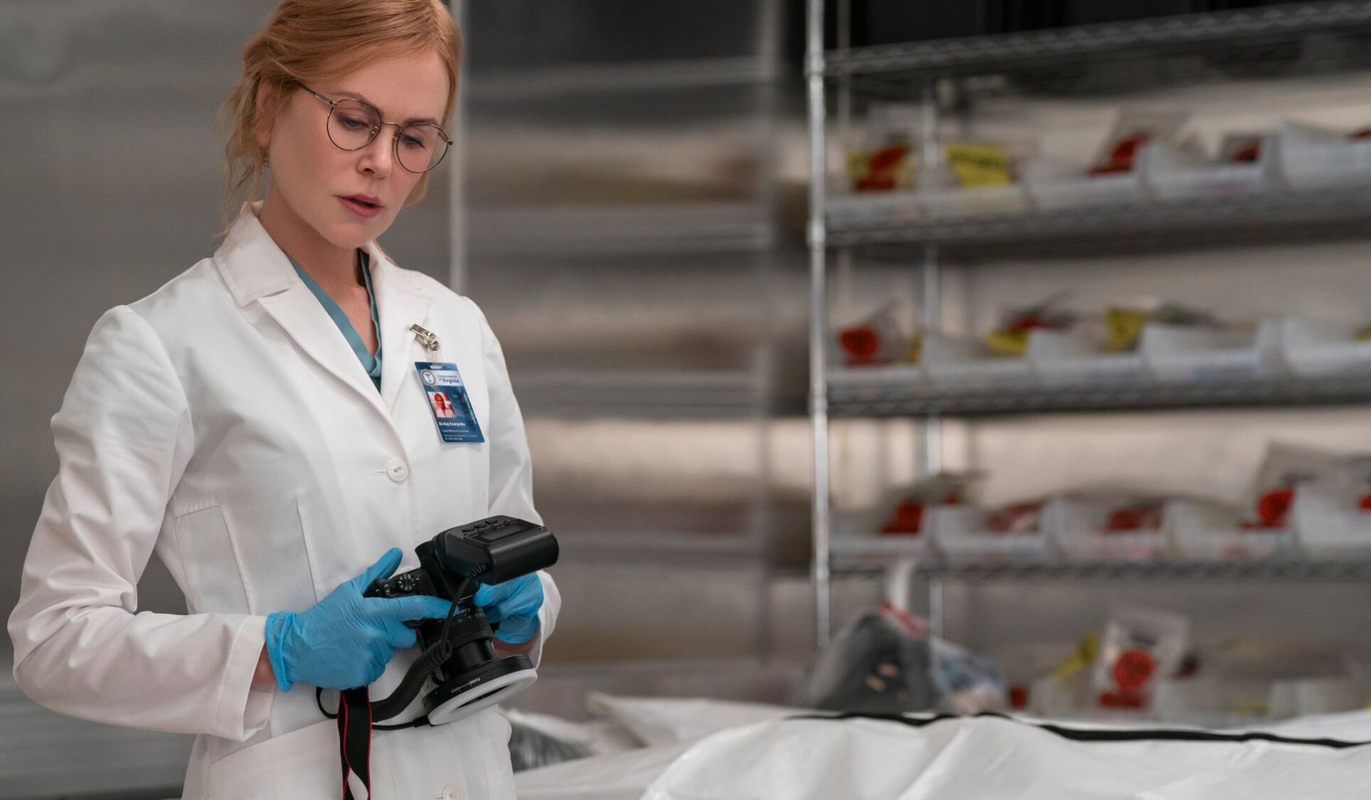 A woman in a white lab coat and blue gloves examines a camera in a laboratory, evoking Scarpetta as she investigates clues in this murder thriller. Shelves with labelled evidence bags are visible behind her, Nicole Kidman-style poise evident.