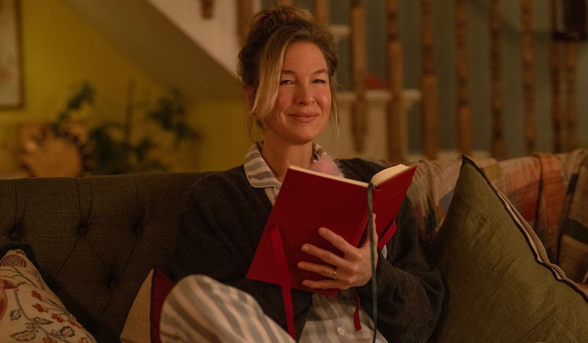 A woman in pyjamas sits on a cosy sofa, smiling as she holds and reads a red diary—perhaps jotting down thoughts after watching her favourite Valentine’s films. The background shows a staircase and warmly lit, homely decor.