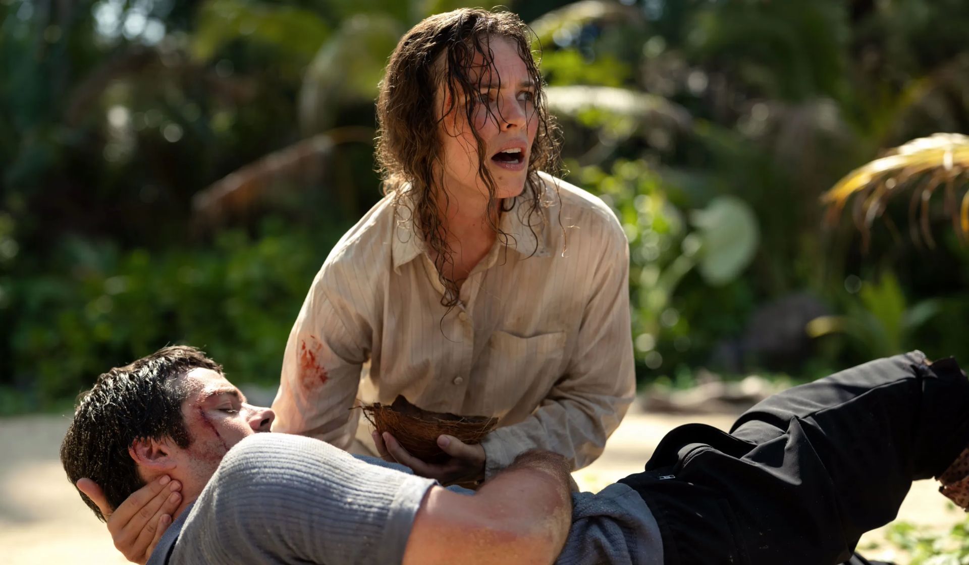 A distressed woman, resembling Rachel McAdams, kneels on sandy ground cradling an injured man. Wet hair frames her face as she silently pleads, “Send Help.” Lush green foliage hints at a tropical backdrop in this comedy thriller scene.