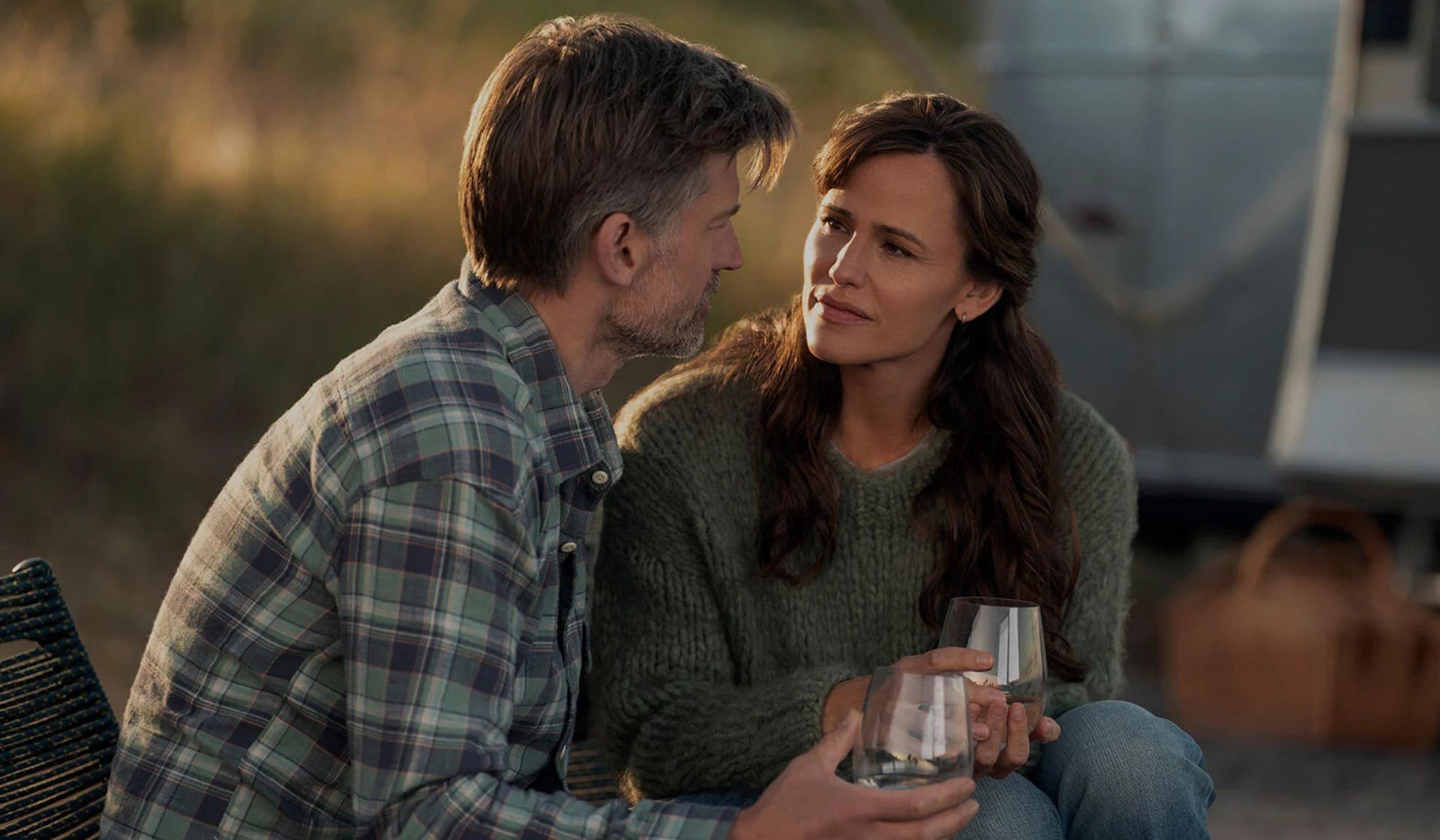 A man and woman sit close together outdoors, holding glasses and looking at each other. The softly blurred background with a picnic basket and camper evokes a relaxed, intimate moment—much like a scene from The Last Thing He Told Me Series 2.