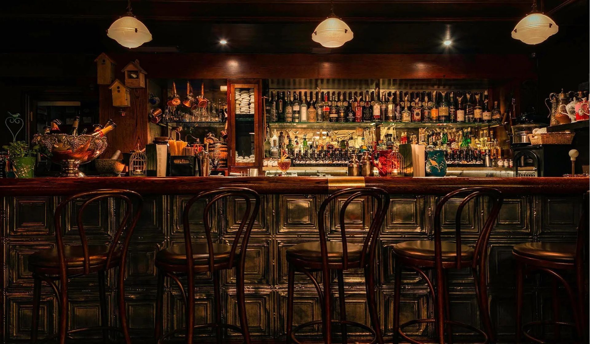 A dimly lit bar reminiscent of classic London jazz bars, with four empty wooden stools at a polished counter, shelves behind stocked with various bottles, glassware, and decorations under warm hanging lights.