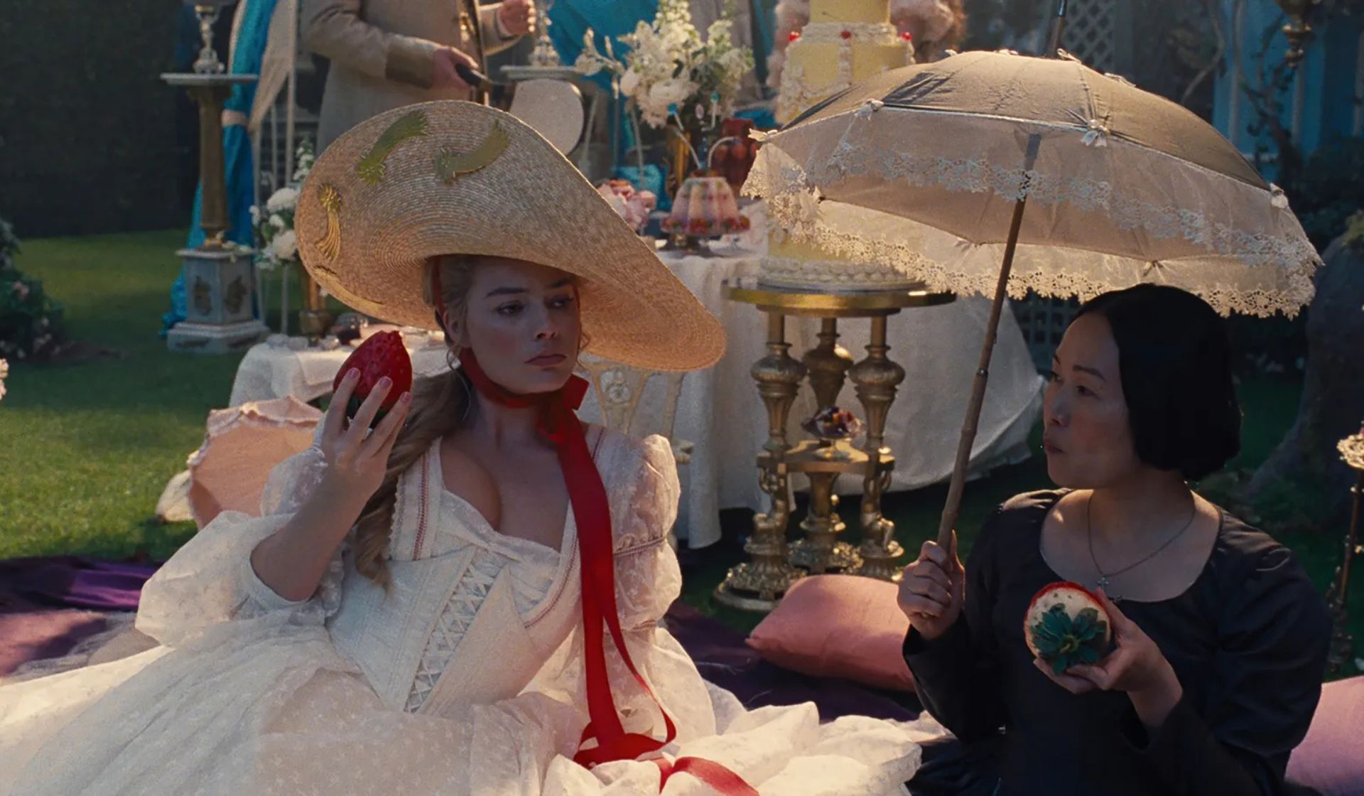 Two women sit at a lavish outdoor tea party, evoking a Wuthering Heights atmosphere. One, in a white dress and hat, holds a red apple; the other, in dark attire, holds an embroidered hoop and lace parasol. Decorated tables and food grace the background.