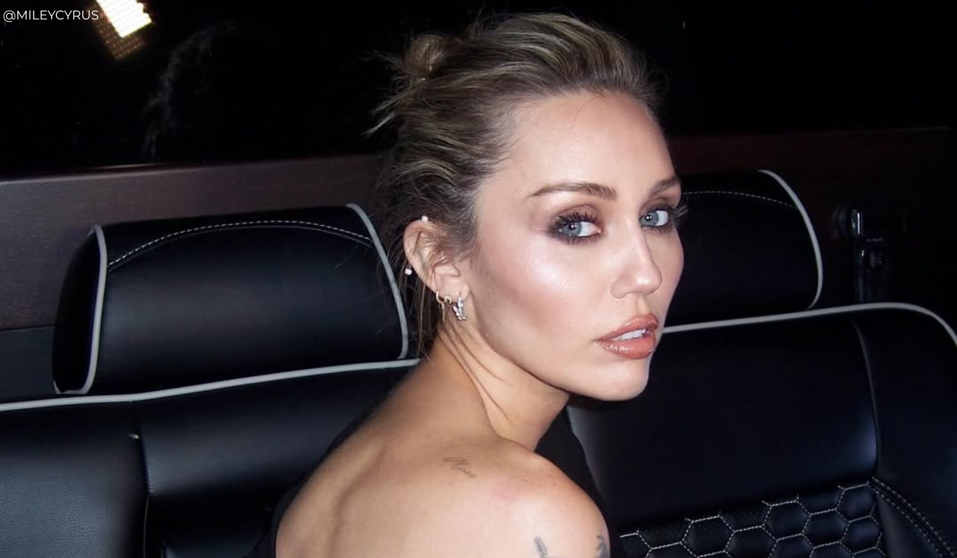 A woman with light skin and blue eyes sits in a black leather car seat, looking over her shoulder at the camera, reminiscent of a Miley Cyrus premiere entrance. Her hair is in a messy bun and she wears subtle make-up with nude lipstick.