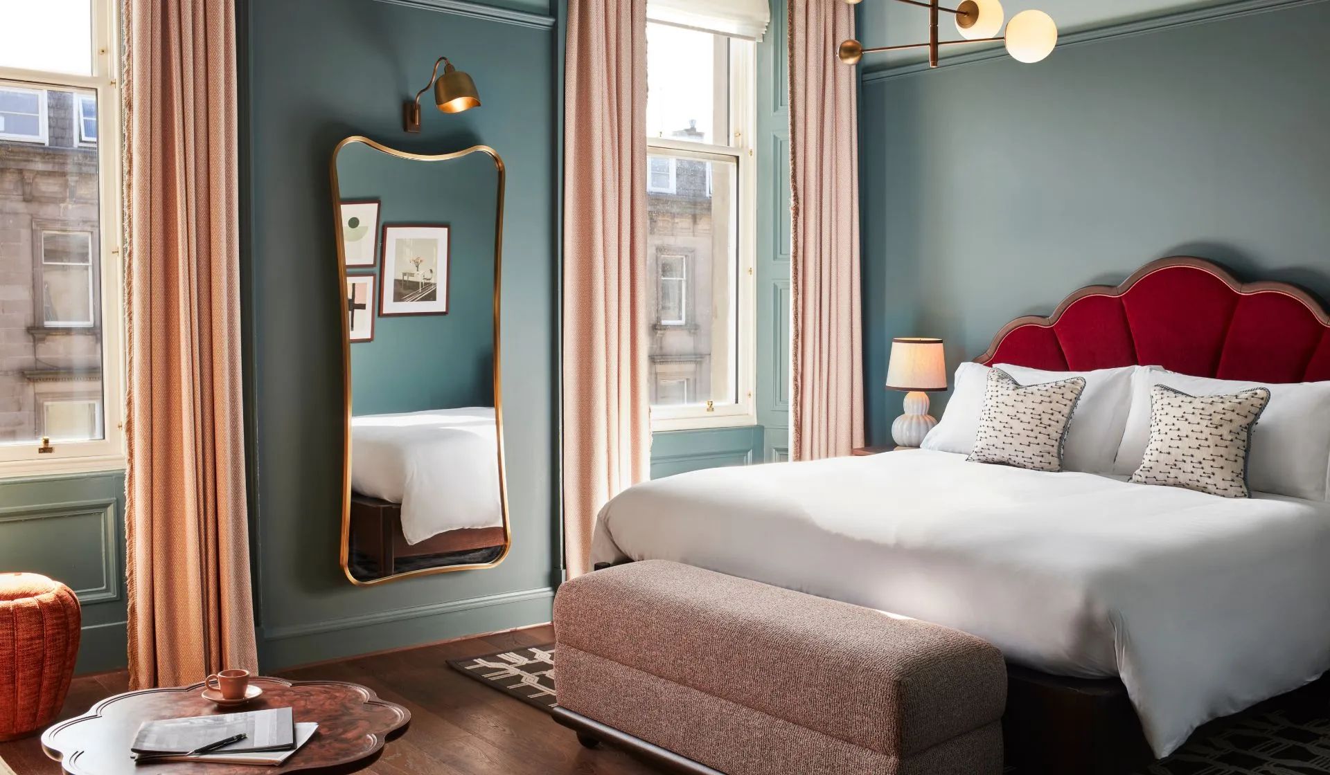 A stylish bedroom inspired by an Edinburgh Insider’s Guide, featuring blue-green walls, a large bed with a red headboard, a bench at the foot, a gold-framed mirror, pink curtains, and soft lighting from lamps and modern light fittings.