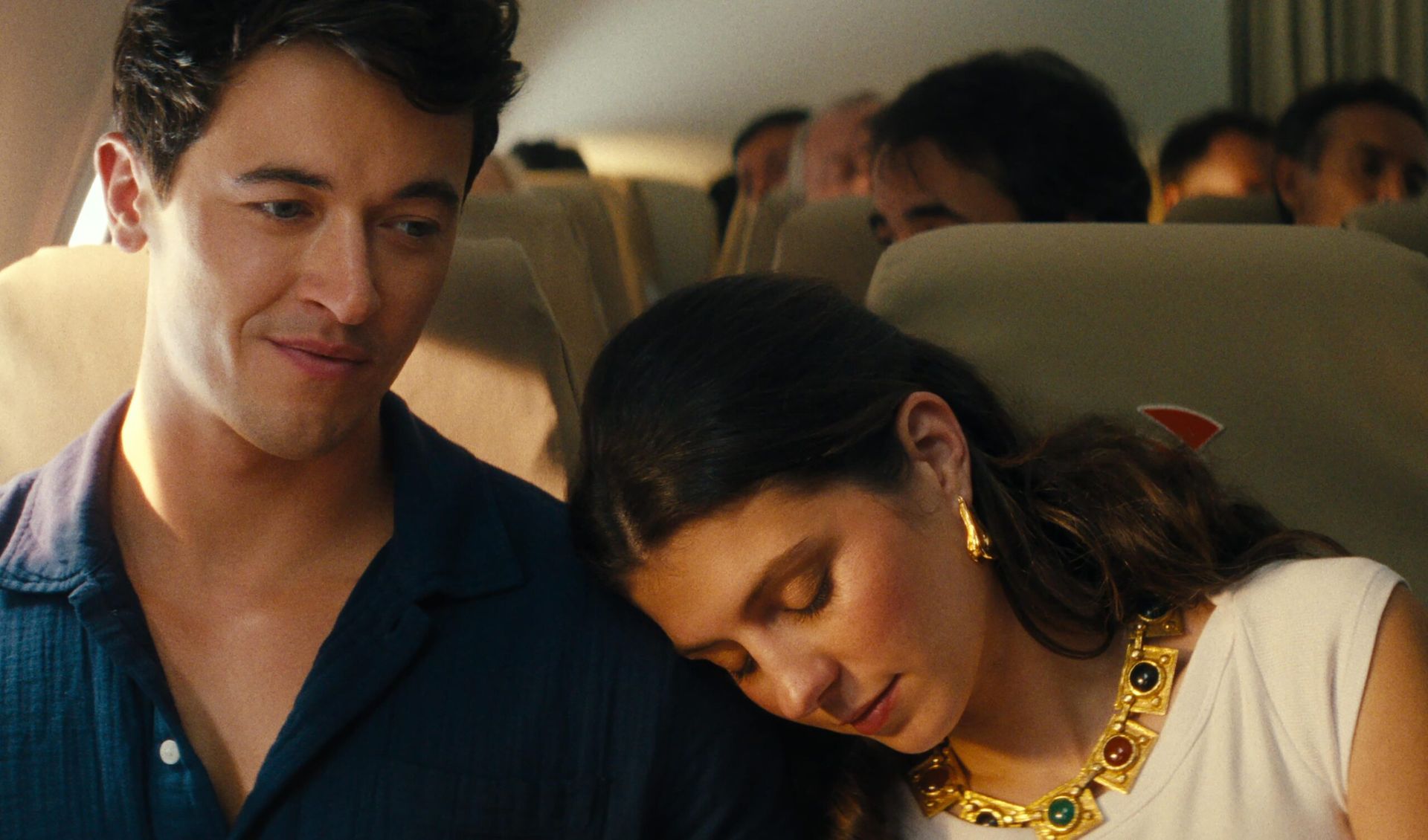 A young woman with long brown hair and gold jewellery rests her head on a young man’s shoulder whilst sitting on an aeroplane, creating a moment reminiscent of valentine’s films. The man smiles gently as other passengers are seated in the background.