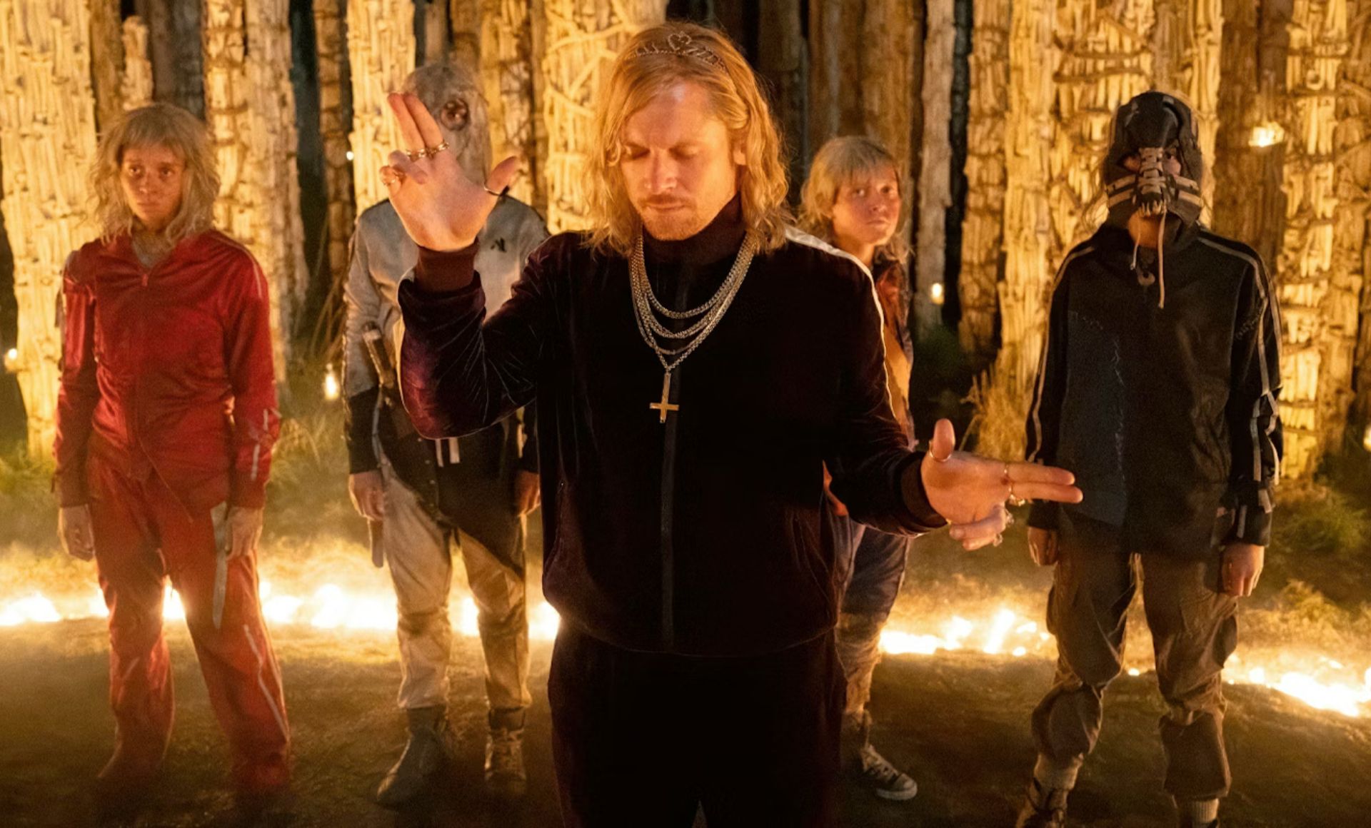 Five people stand in a forest circle lit by candles at night. The central figure wears gold chains and raises both hands, whilst the others, dressed in distinctive outfits, evoke the mystery of Upcoming Films 2026 amid tall, textured tree trunks.