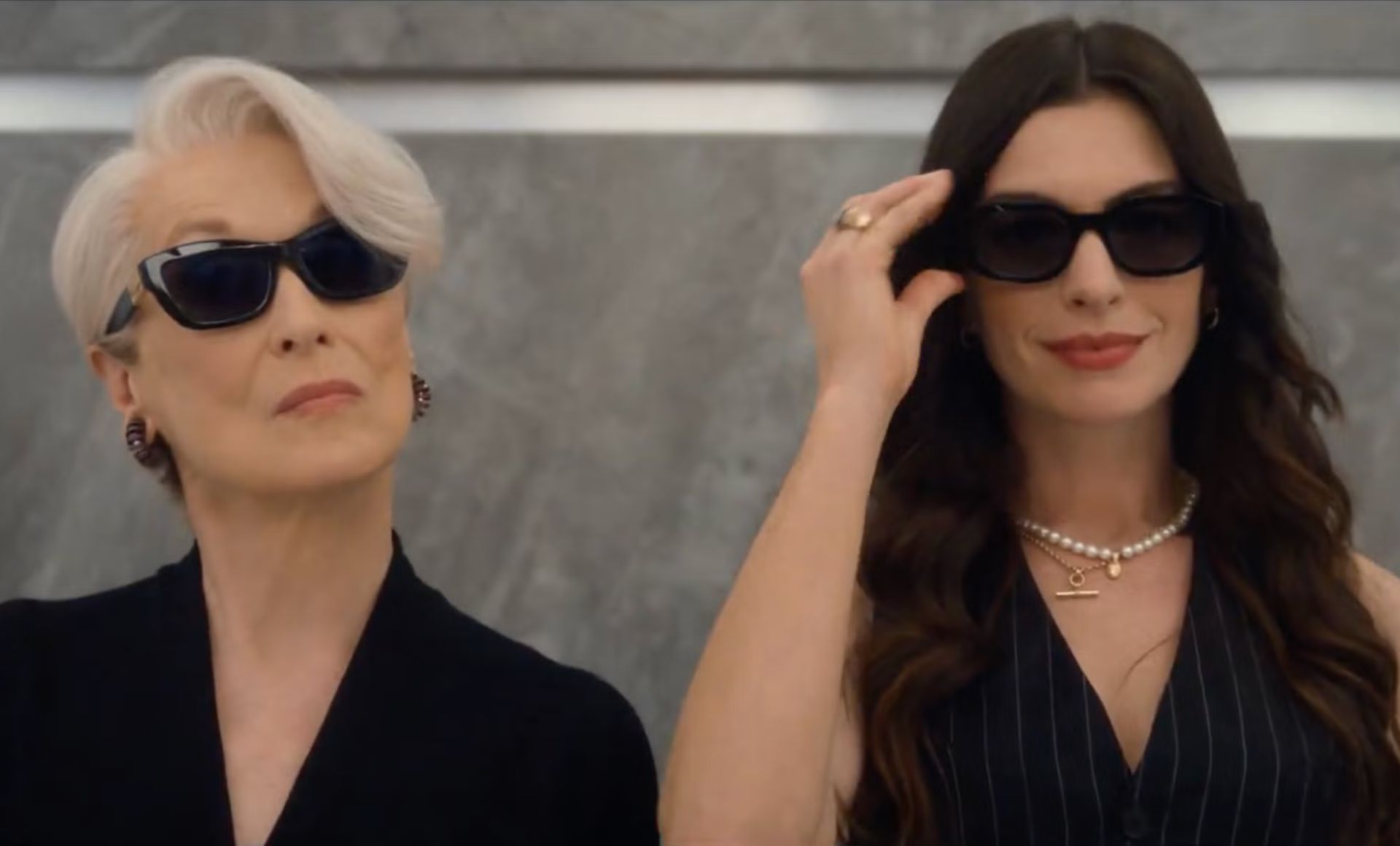 Two women wearing dark sunglasses and elegant black outfits stand side by side against a marble background, exuding confidence and style—like true film stars 2026. One has short white hair, the other long brown hair, both embodying cinematic allure.