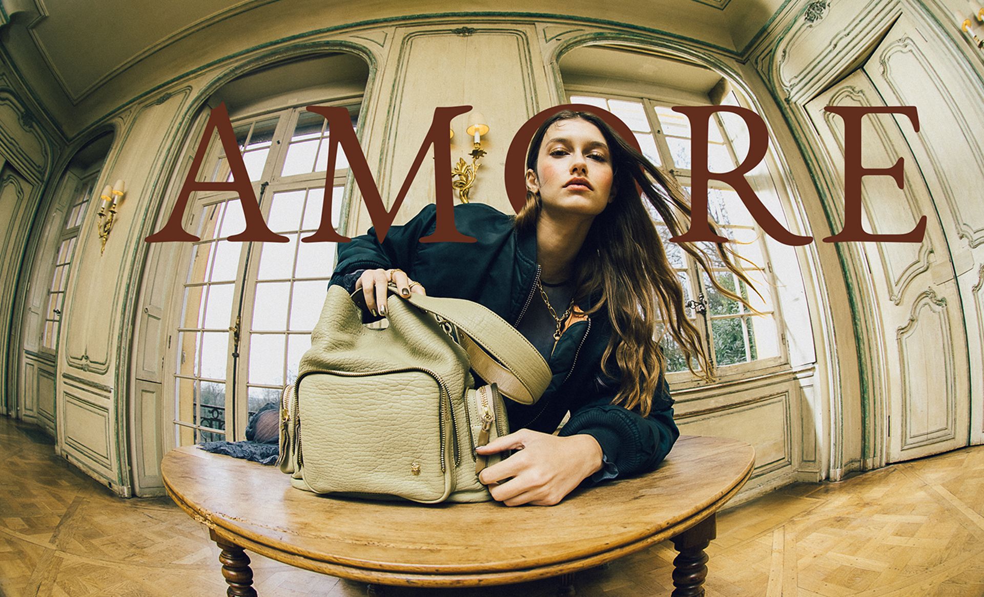 A woman sits at a small round table with a beige bag from one of today’s chic small fashion brands, posing confidently in an ornate room. Large text reading AMORE is overlaid across the image.