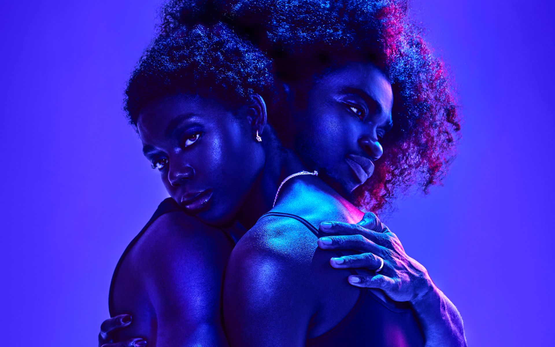 Two women with natural curly hair embrace under dramatic purple and blue lighting against a soft background, creating a bold, intimate portrait perfect for a weekend guide seeking inspiring visual stories.