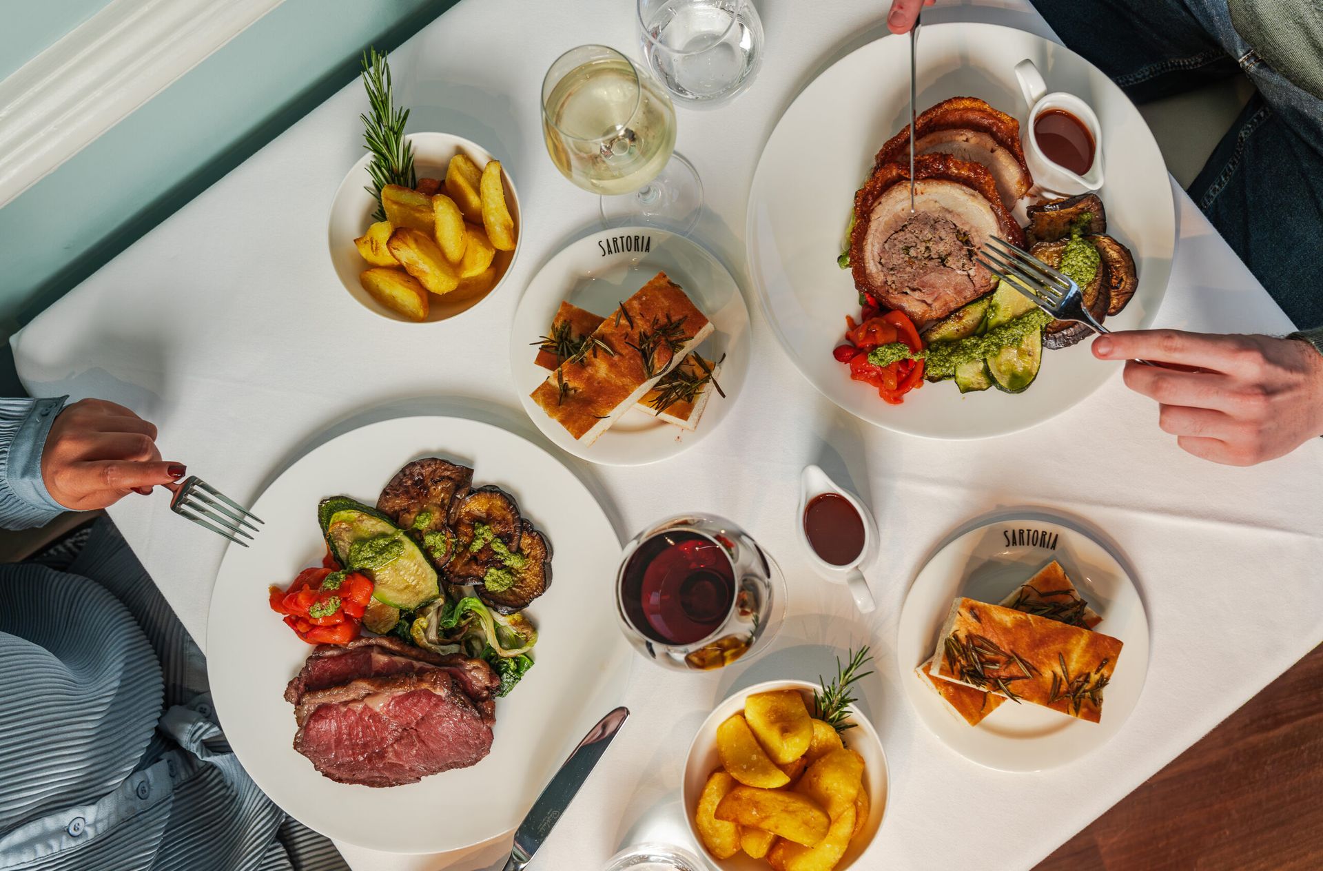 A table set for two with plates of roast beef, rolled pork, roast potatoes, vegetables, glasses of red and white wine, and slices of focaccia bread on a white tablecloth—perfect for your weekend guide to cosy dining.