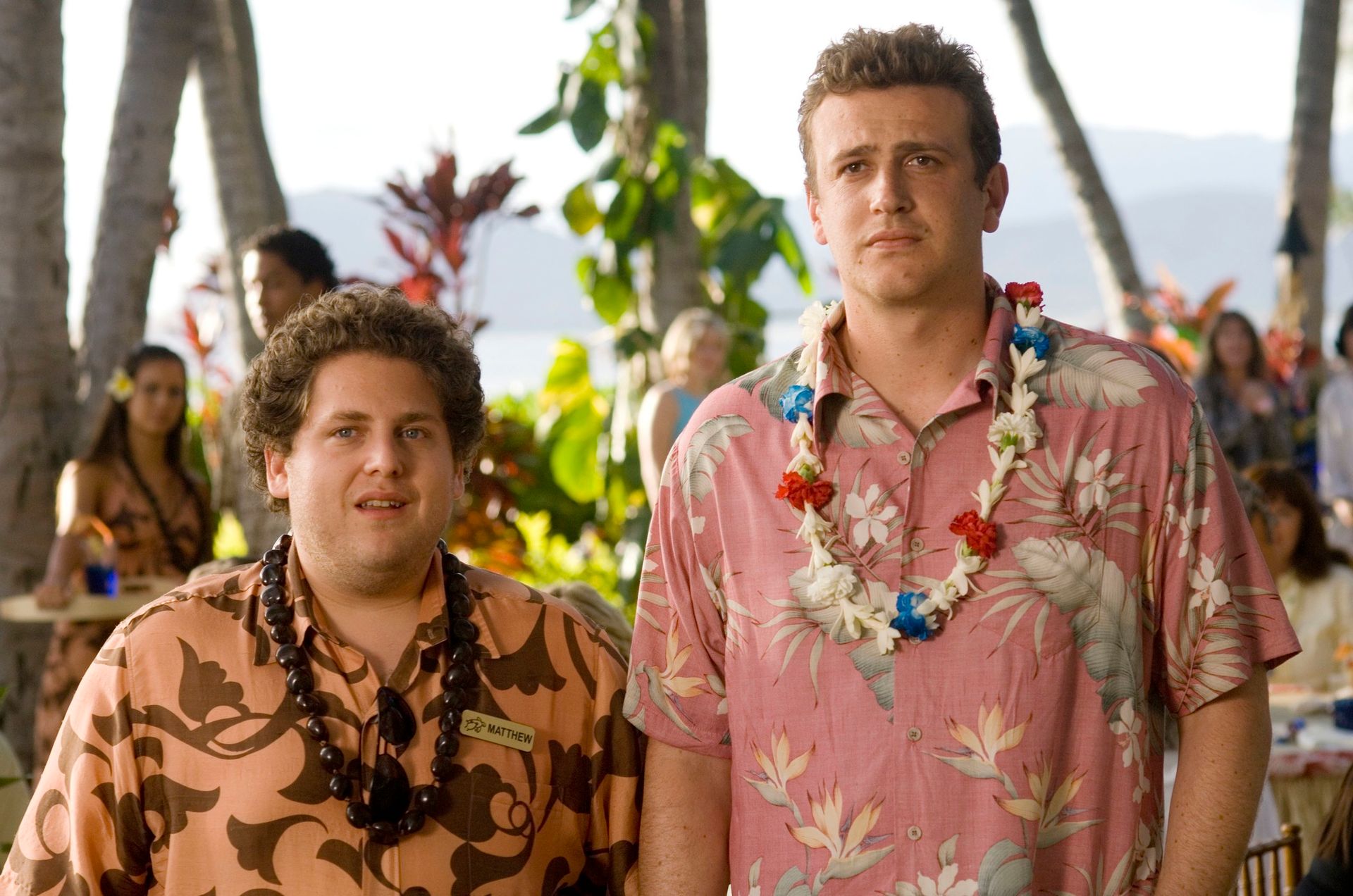Two men stand side by side outdoors in a tropical setting, both wearing Hawaiian shirts and leis. From a bloke’s perspective, it’s like a scene straight out of the best romantic comedies: palm trees, plants, and a relaxed beachside vibe fill the background.