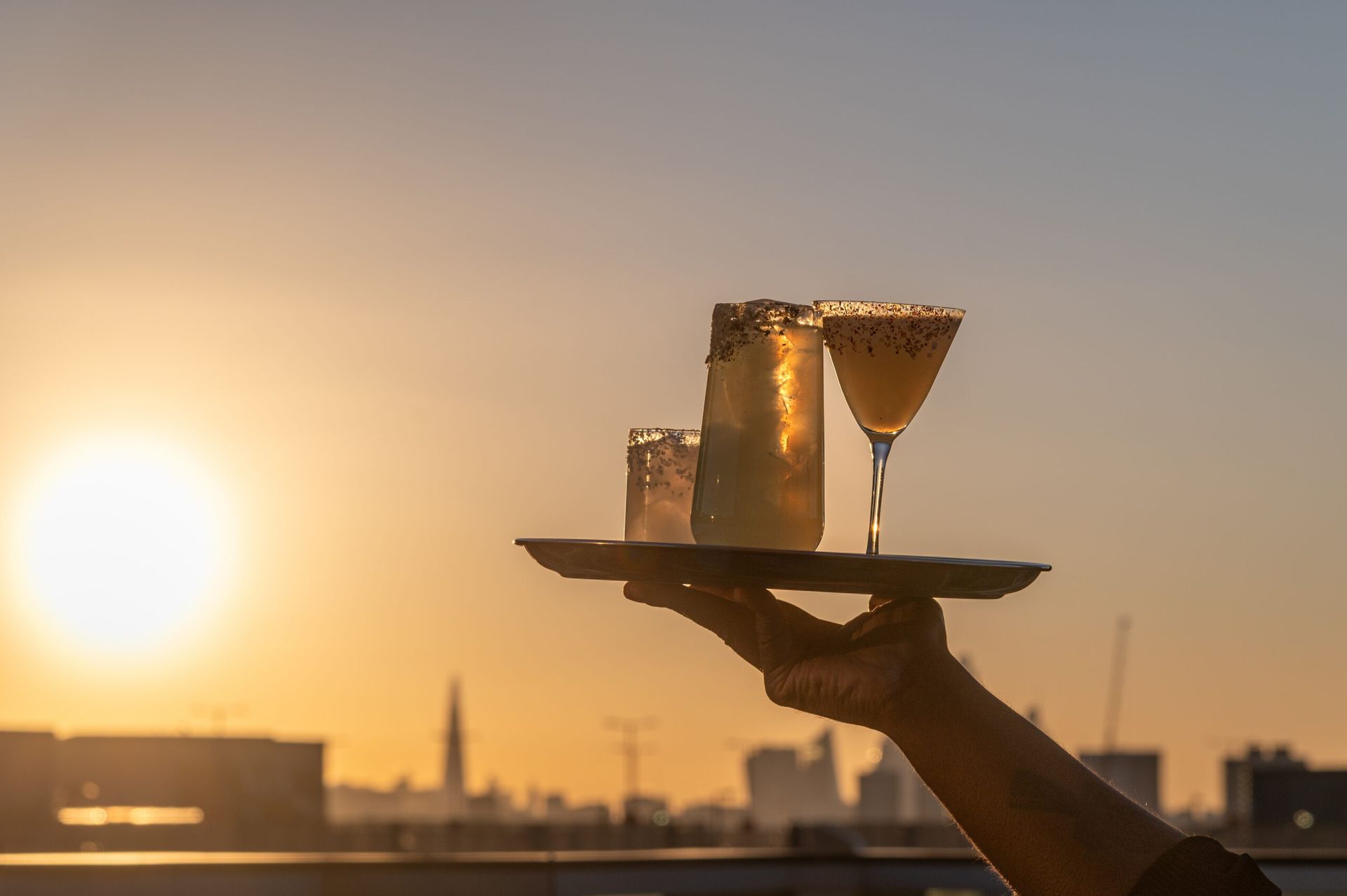 A hand holds up a tray with three cocktails against a city skyline at sunset, the sun glowing brightly in the sky—perfect inspiration for your Weekend Guide.