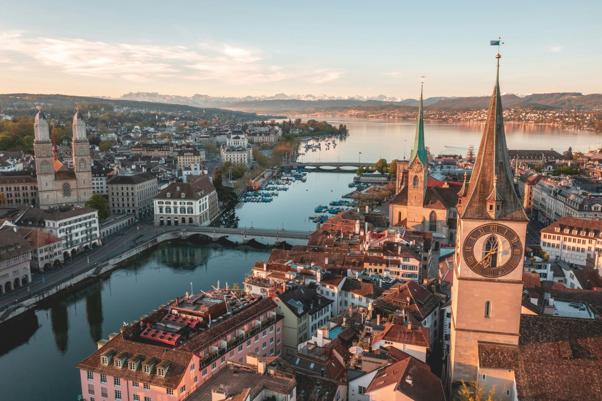 Aerial view of Zurich, Switzerland, with historic buildings, church towers, the Limmat River, and the iconic fairytale hotel Dolder Grand set against distant mountains under a partly cloudy sky at sunset.