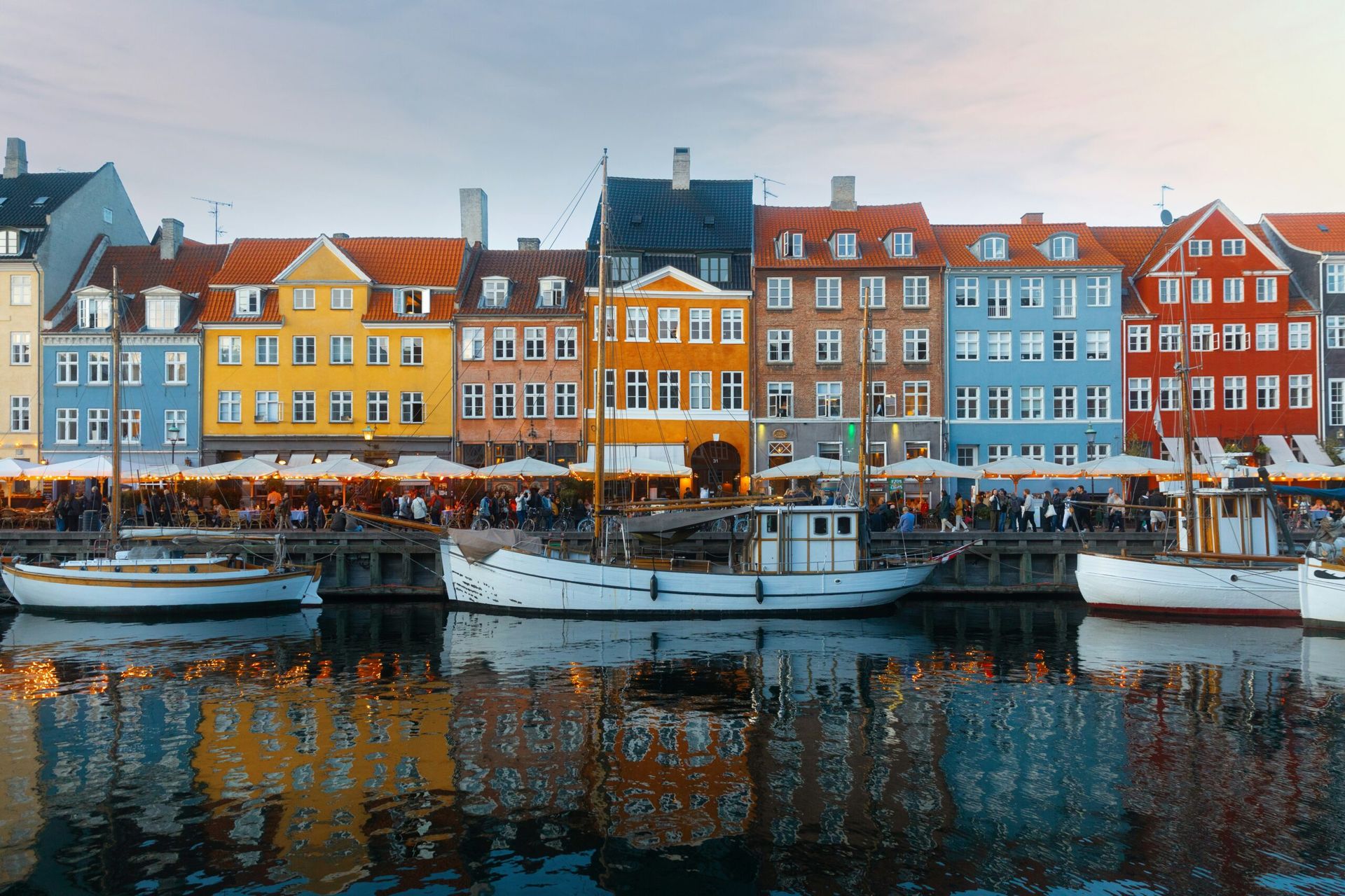 Colourful buildings line the Nyhavn waterfront in Copenhagen, with docked boats reflected in the calm water and crowds at outdoor cafés—an inviting scene for your 2026 travel adventure, featuring highlights like Villa Copenhagen.