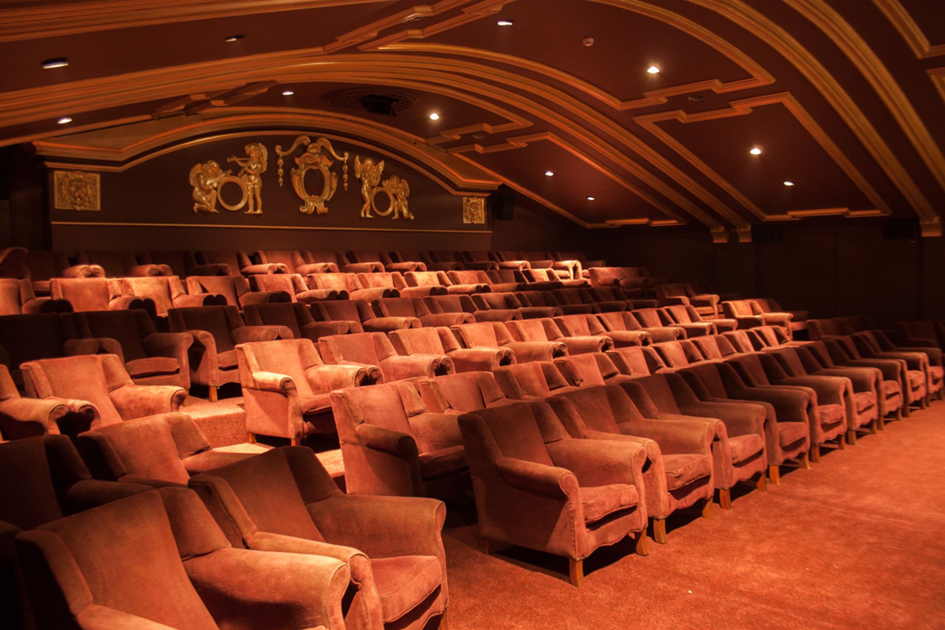 An empty, ornate theatre reminiscent of London independent cinemas, with rows of plush, reddish-brown armchairs, decorative gold ceiling accents, and warm lighting creating an inviting atmosphere.