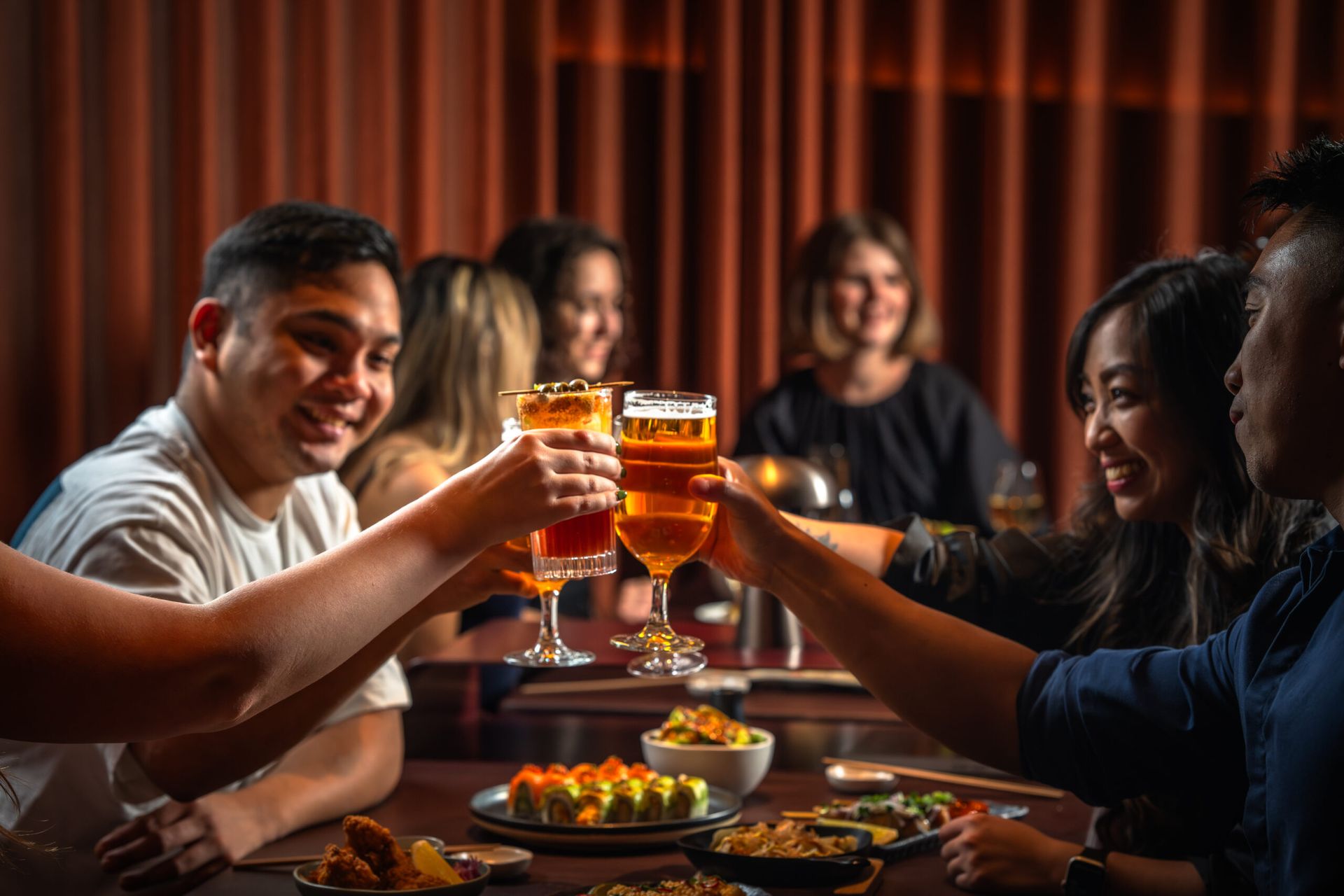 A group of friends sit around a table clinking glasses of beer and cocktails, smiling and enjoying a meal with plates of food in a warm, dimly lit restaurant—perfect for fighting the January blues on a cosy weekend.