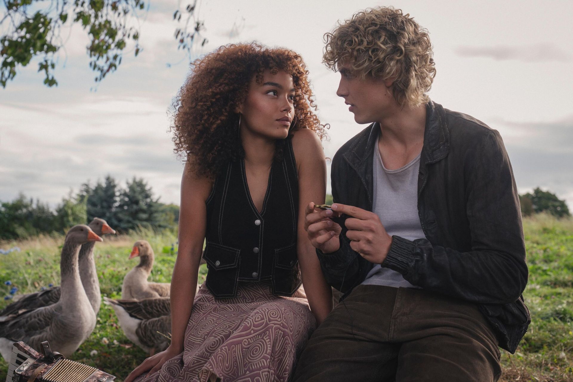 A young woman and man sit close together outdoors, looking at each other. The man holds a small object. Three geese and trees enhance the serene setting, like a scene from the biggest films 2026 or featuring stars to watch 2026.