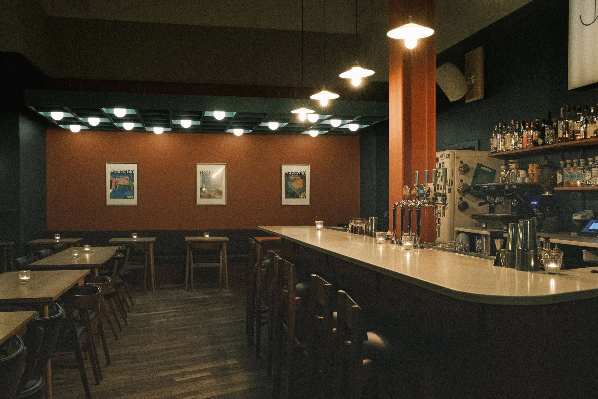 A dimly lit bar featured in the Manchester Guide, with empty wooden tables and chairs, a long counter with barstools, several taps, and shelves with bottles. Framed art hangs on a rust-coloured wall as pendant lights illuminate the cosy space.