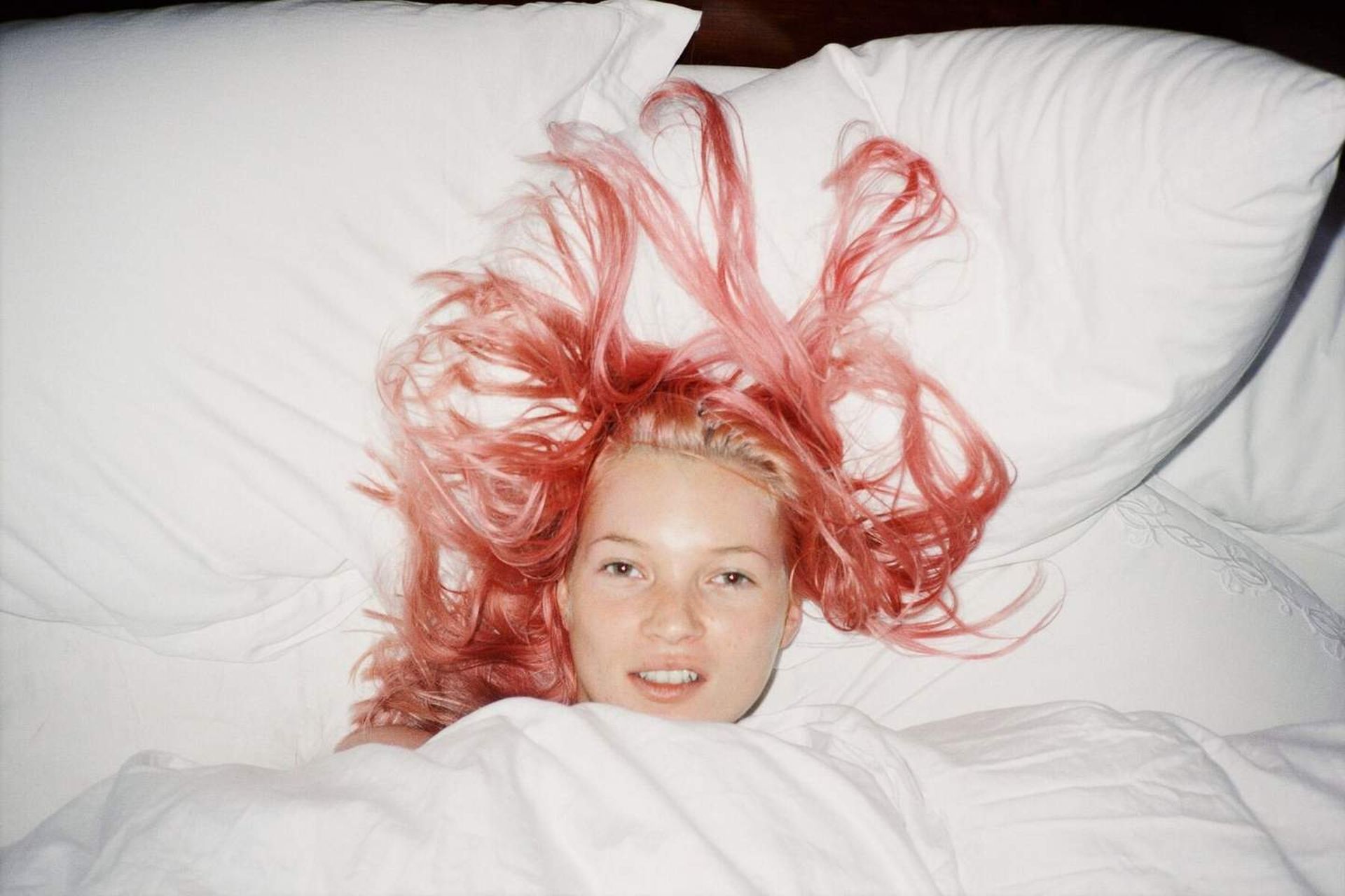 A person with pink hair lies in bed, covered up to their shoulders with a white duvet. Their hair fans out on the white pillow, creating a striking visual effect reminiscent of looks seen at top fashion shows. They are looking towards the camera.