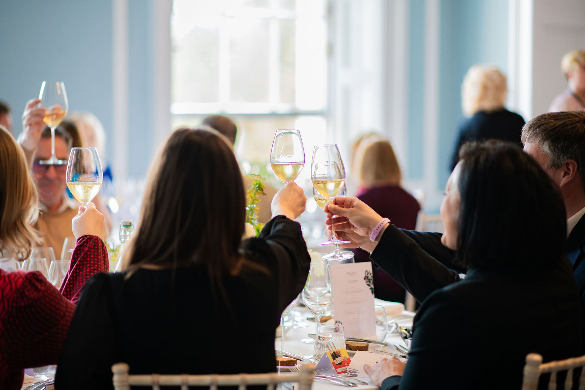 People seated around a table raise wine glasses in a toast at a formal gathering, with bright natural light coming through large windows—a perfect scene for any London weekend guide.