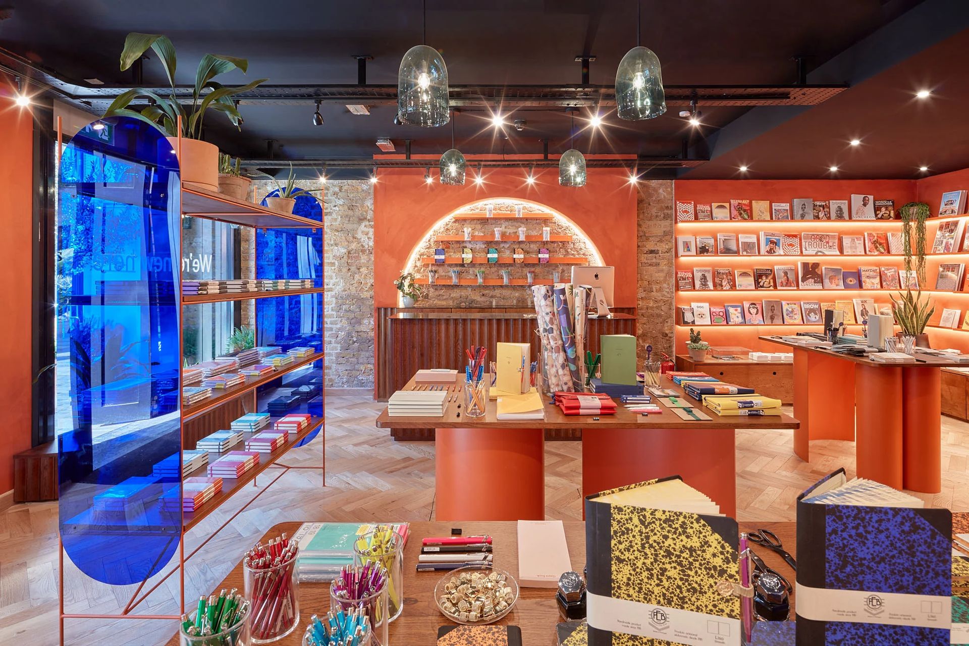 A vibrant stationery shop with orange walls, wooden floors, shelves of books and notebooks, colourful pens on tables, houseplants, and a blue glass divider. Warm lighting and decorative accents create a cosy atmosphere.
