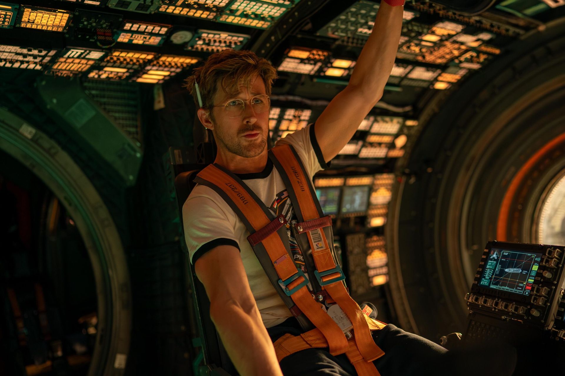 A man with glasses and messy hair, rumoured among the stars to watch 2026, wears a harness and casual clothes as he sits inside a high-tech spacecraft filled with control panels and illuminated buttons, gripping an overhead handle.