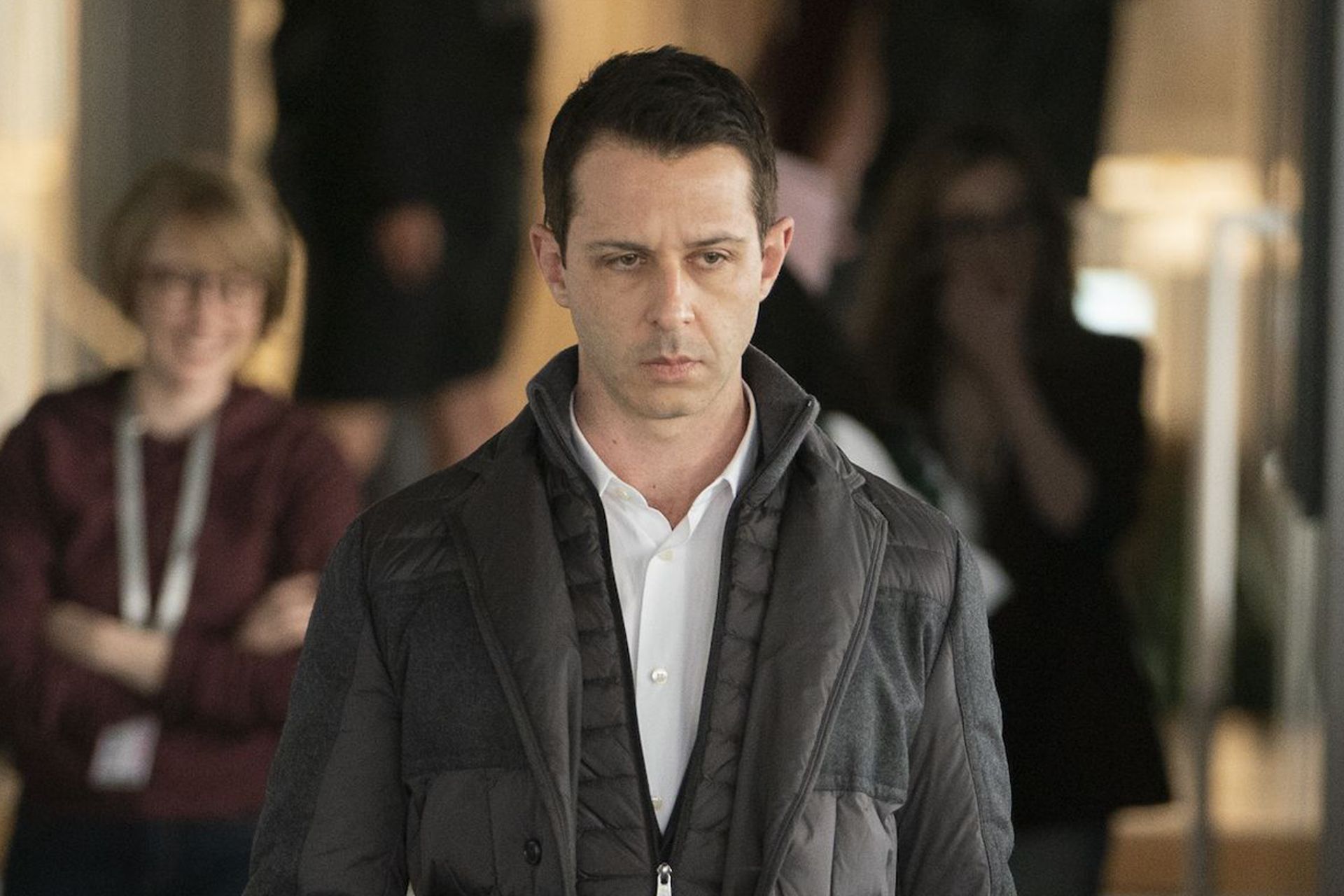A man with short dark hair, wearing a white shirt and a dark quilted jacket, looks serious as he walks indoors. Two blurred people stand in the background—one with arms crossed and smiling, hinting at the Stars to Watch 2026.