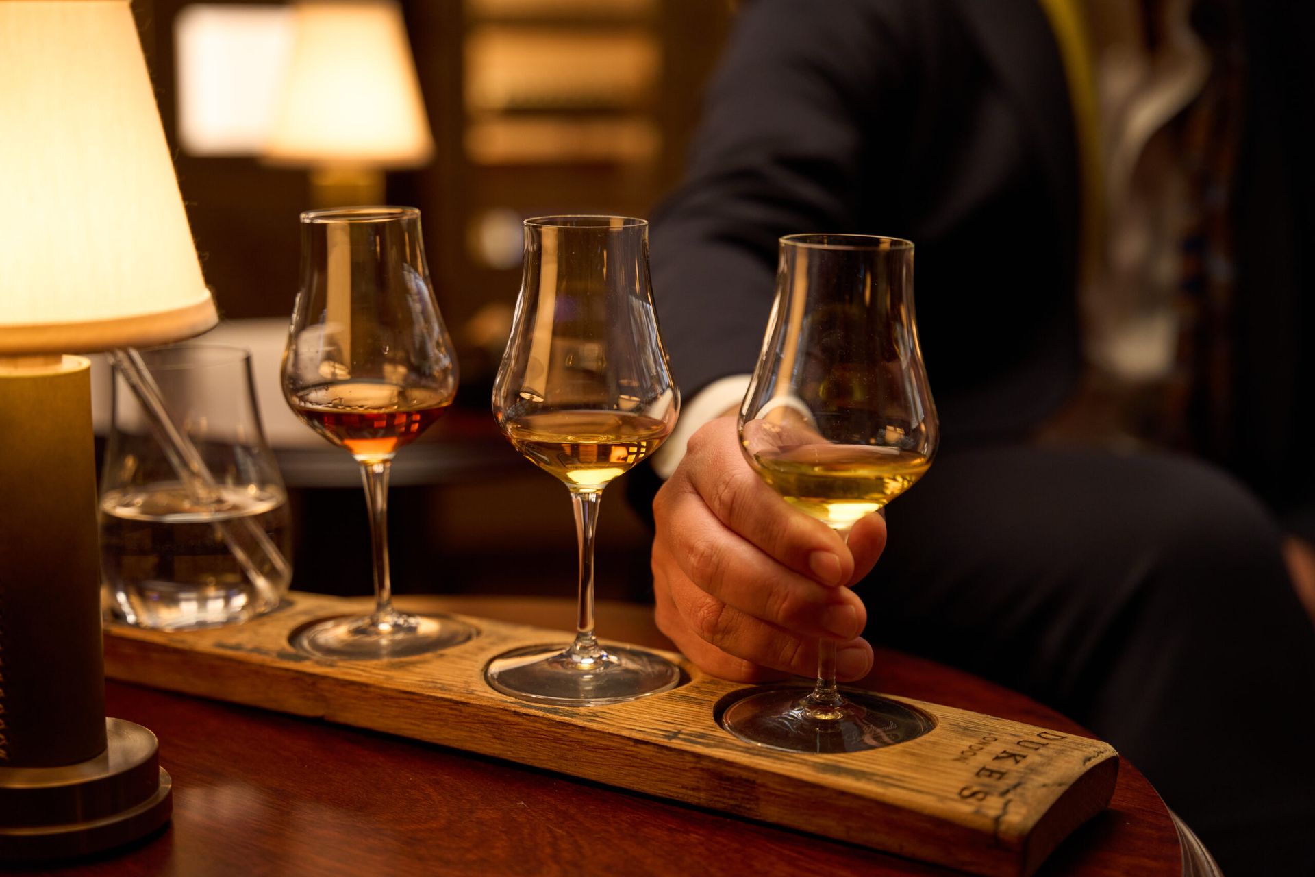 A person in a suit selects a glass of whisky from a tasting flight on a wooden tray, part of exclusive London February pop-ups, with three glasses of different shades set on a polished table beside a lamp and water glass.